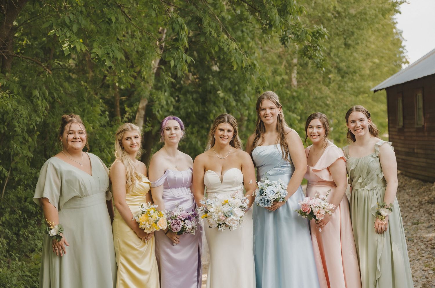 Seven bridesmaids in mismatched pastel gowns holding bouquets pose together at an outdoor wedding.