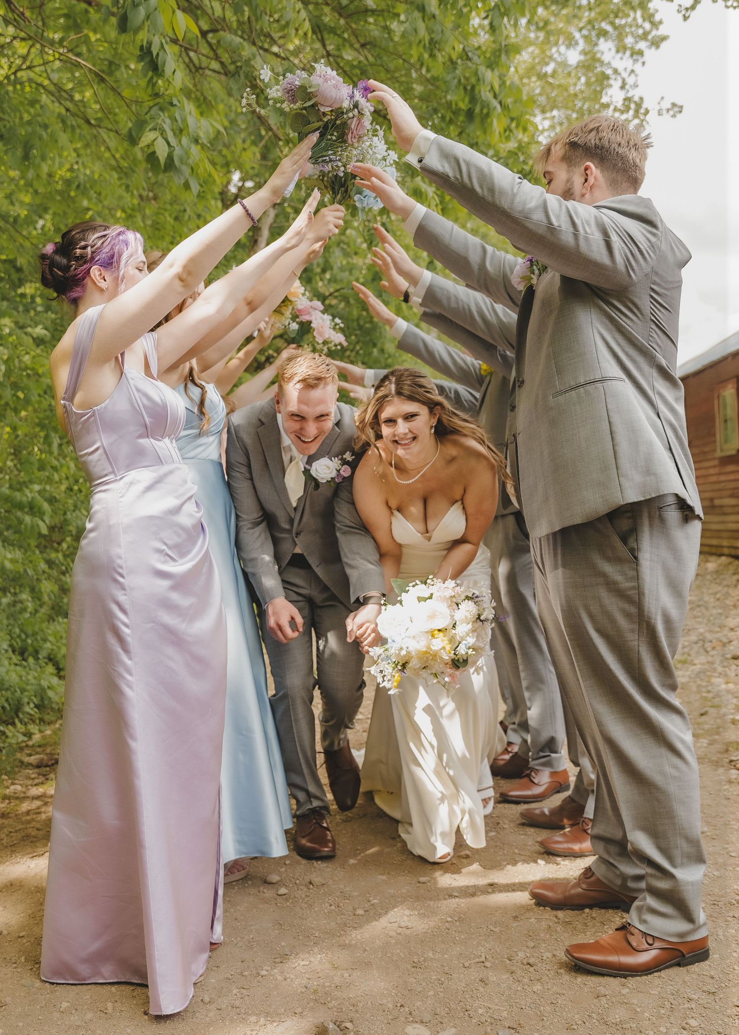 Wedding party forming an arch with bouquets as bride and groom walk beneath in outdoor ceremony.