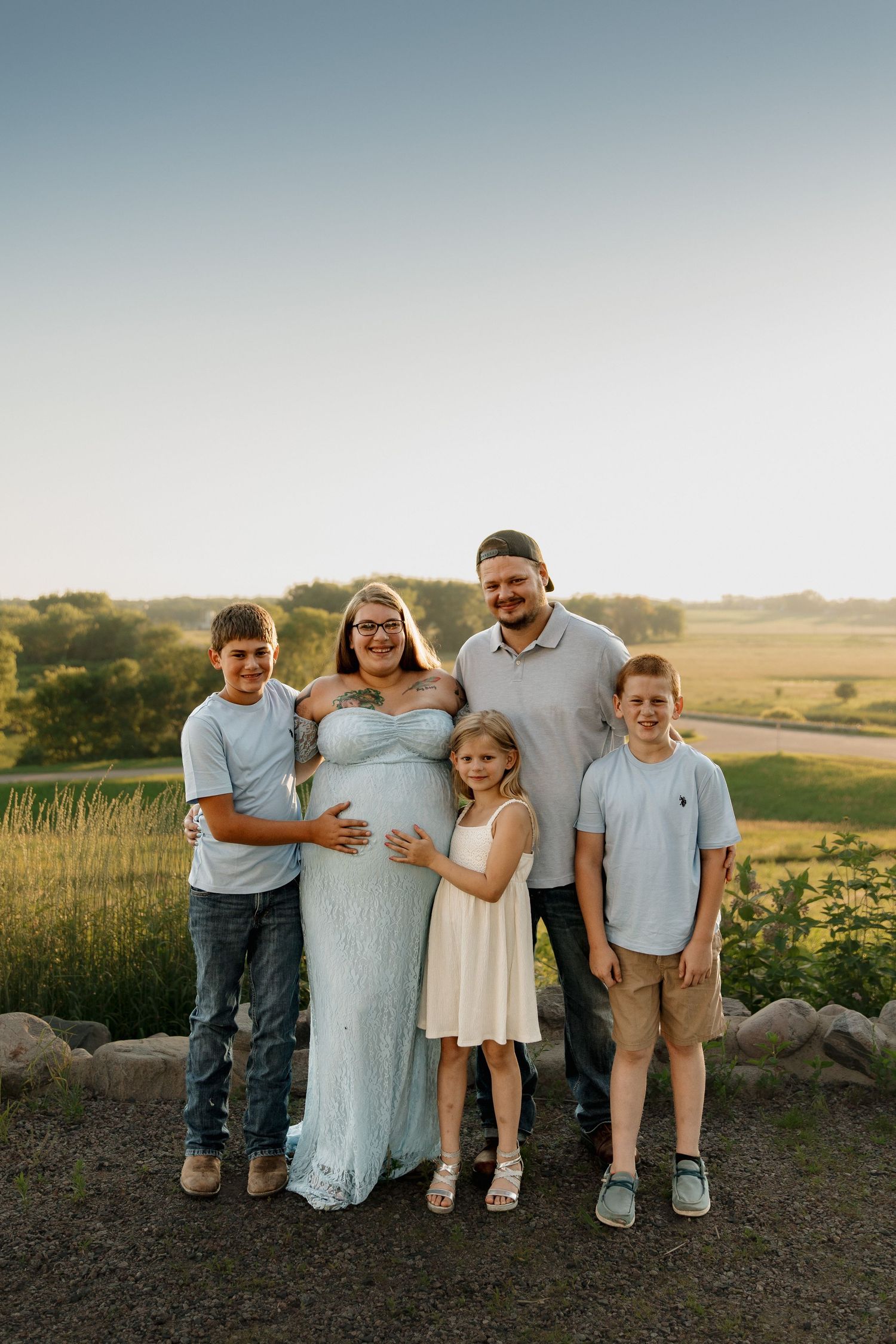 Pregnant woman in light blue gown poses with husband and three children outdoors in a sunny green field.