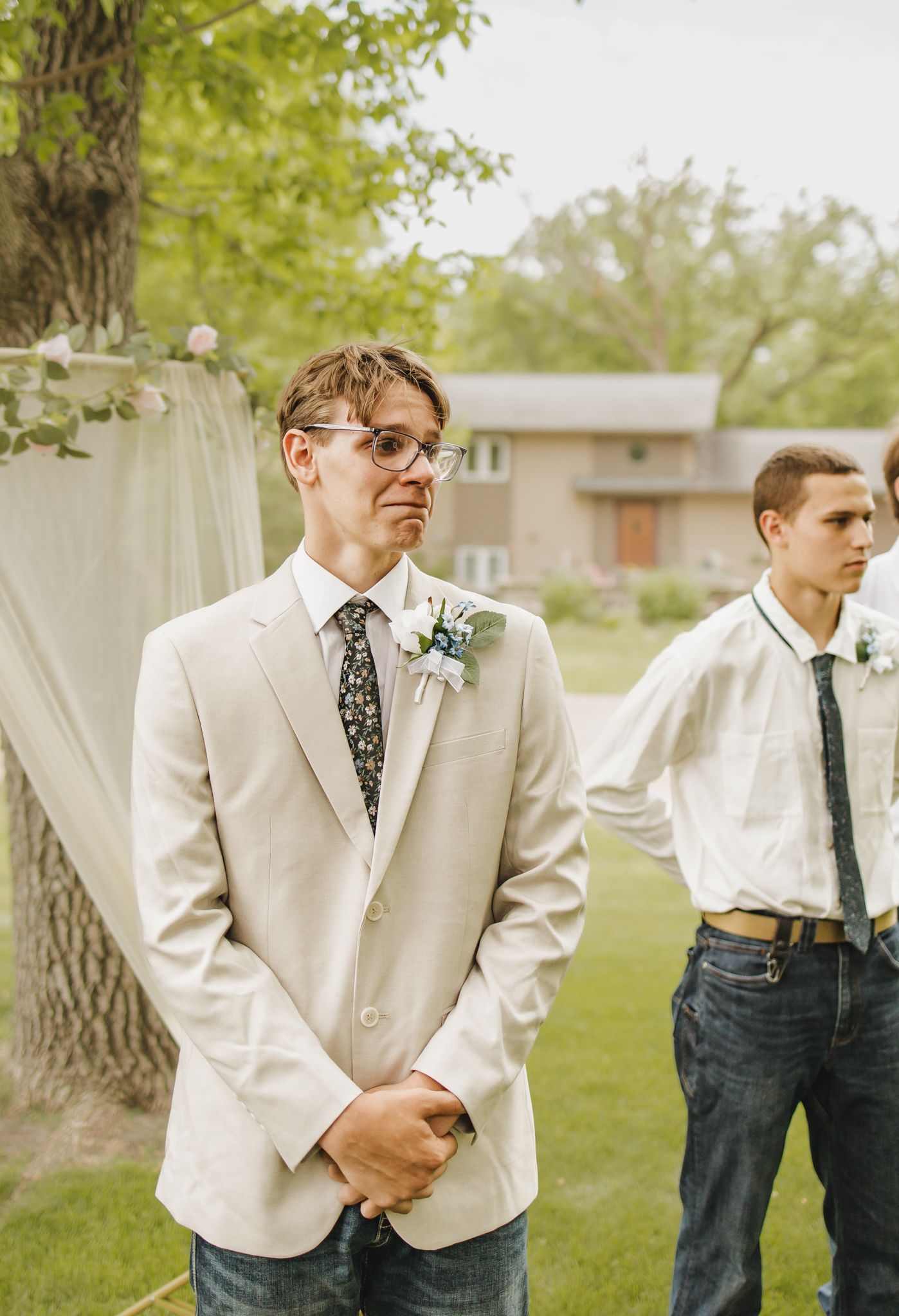 Emotional groom in beige suit with floral boutonniere awaiting bride at outdoor backyard wedding ceremony.