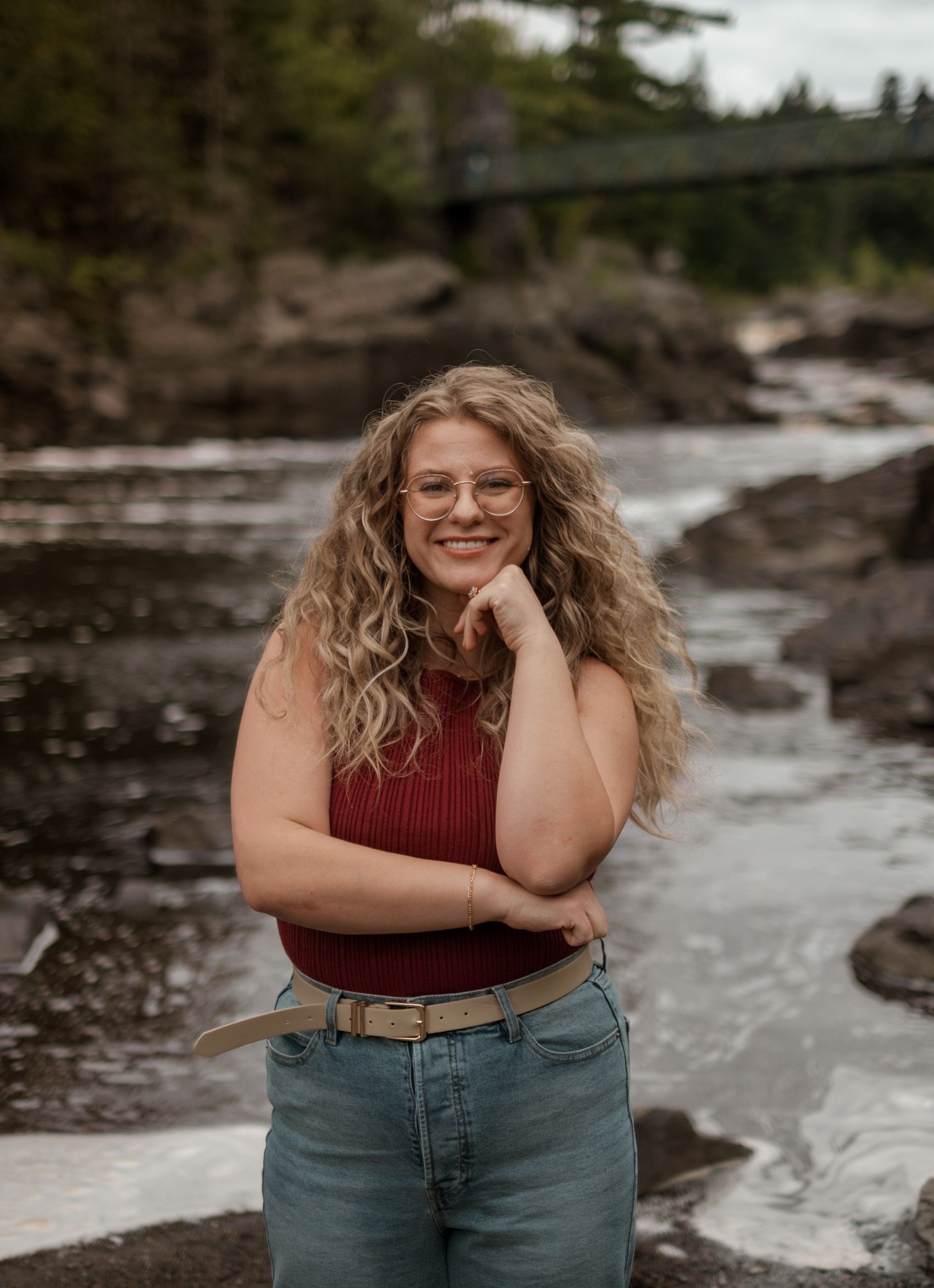 Person in burgundy top and jeans poses near rocky riverbank on cloudy day.