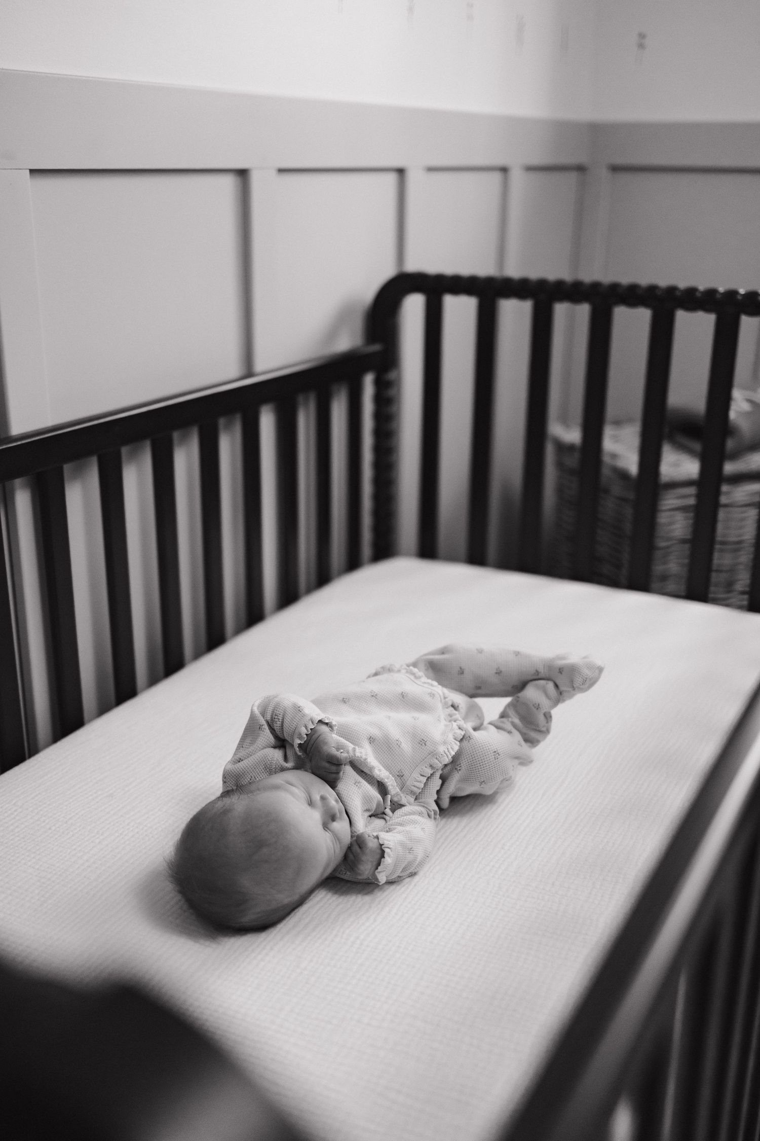 A peaceful newborn baby lies in a dark wooden crib on a white mattress in a black and white photo.