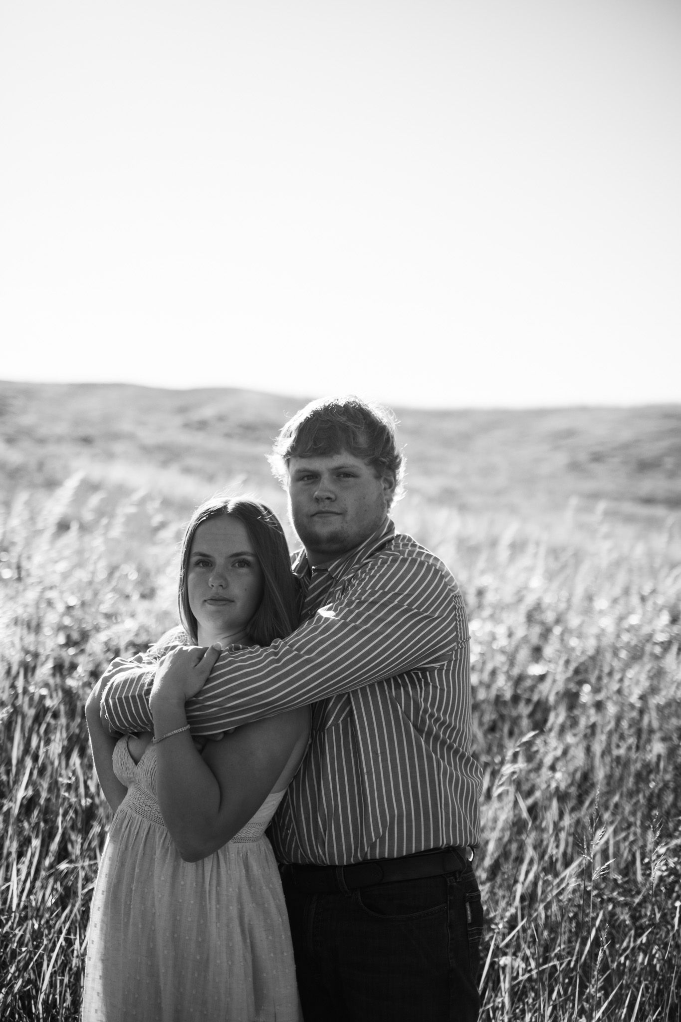 Black and white photo of a young couple embracing in a field, man wrapping arms around woman in a striped shirt.