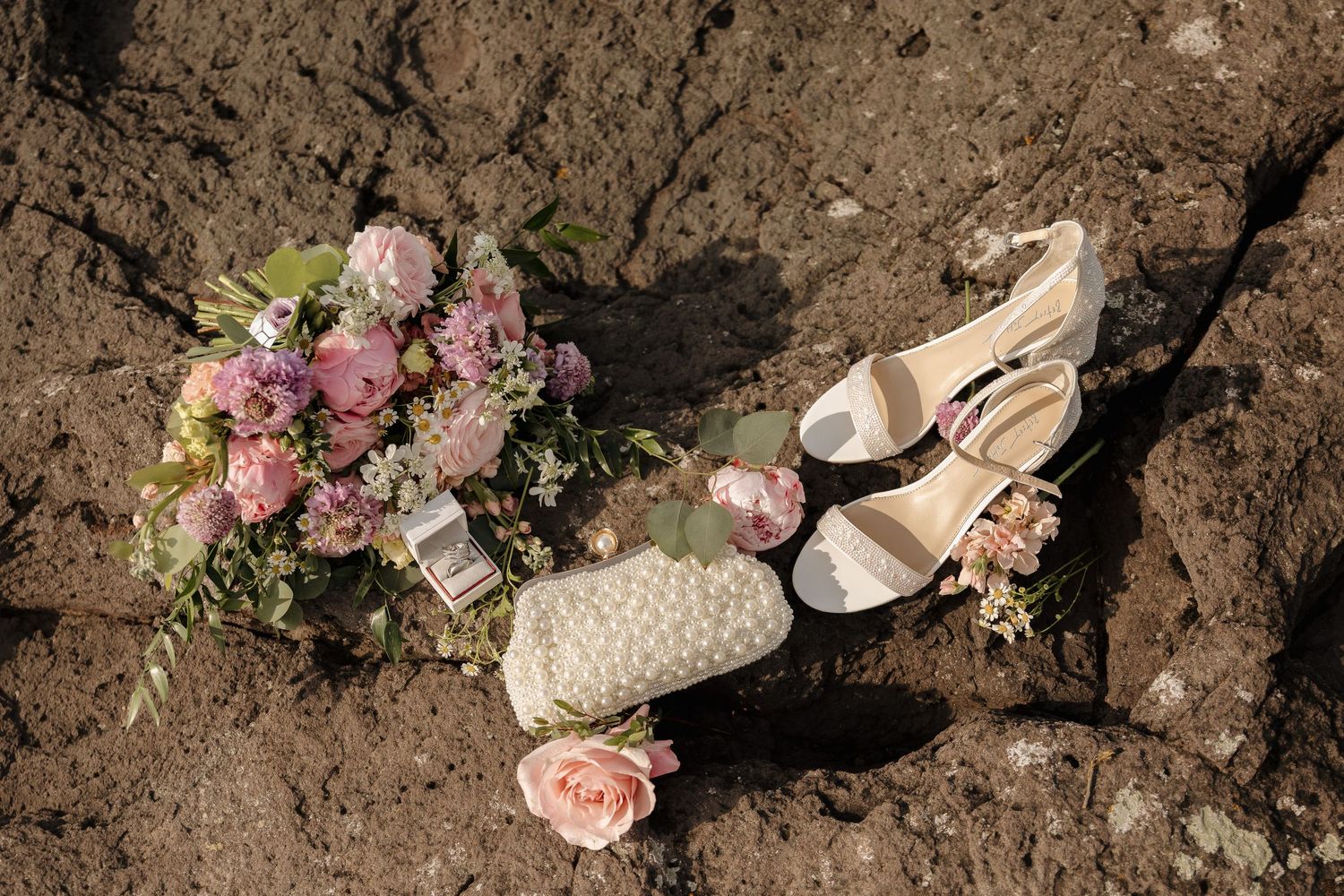 Bridal flat lay with pink flower bouquet, white heeled sandals, pearl clutch, and rose on rocky surface.