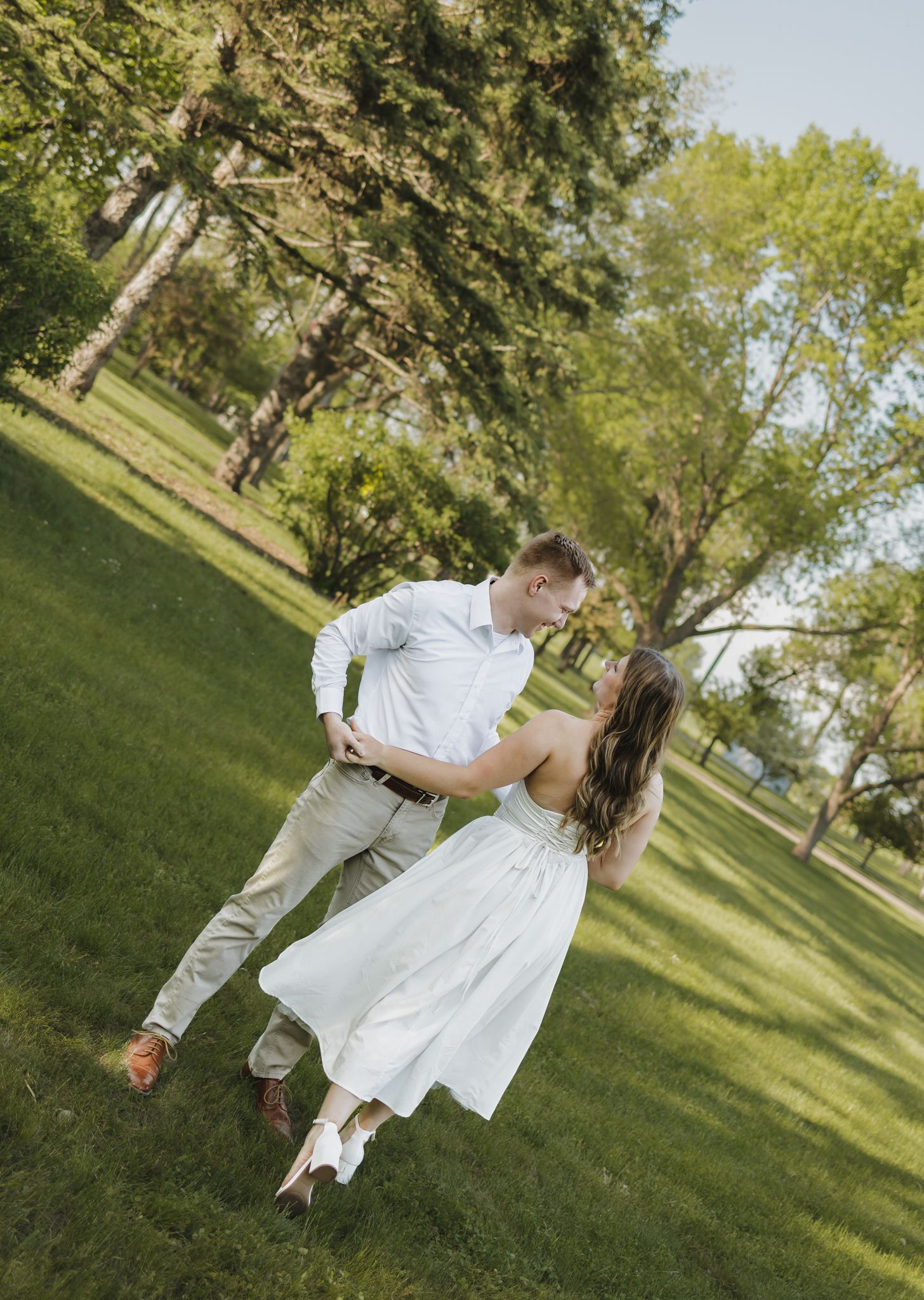 Couple dancing in a lush green park, woman in white dress, man in white shirt and khakis, sharing a romantic dip.