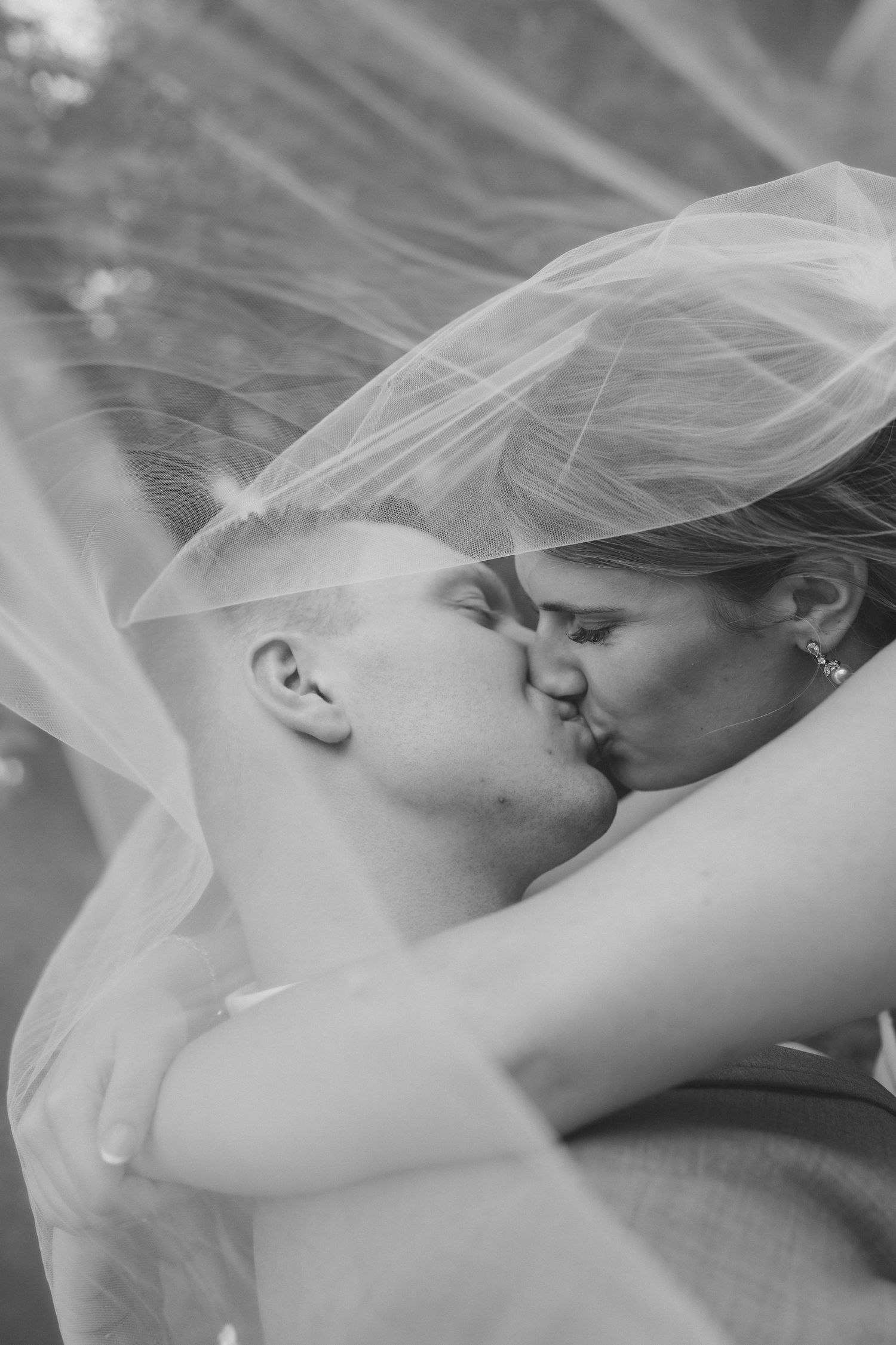 Black and white photo of a bride and groom kissing under a flowing wedding veil outdoors.