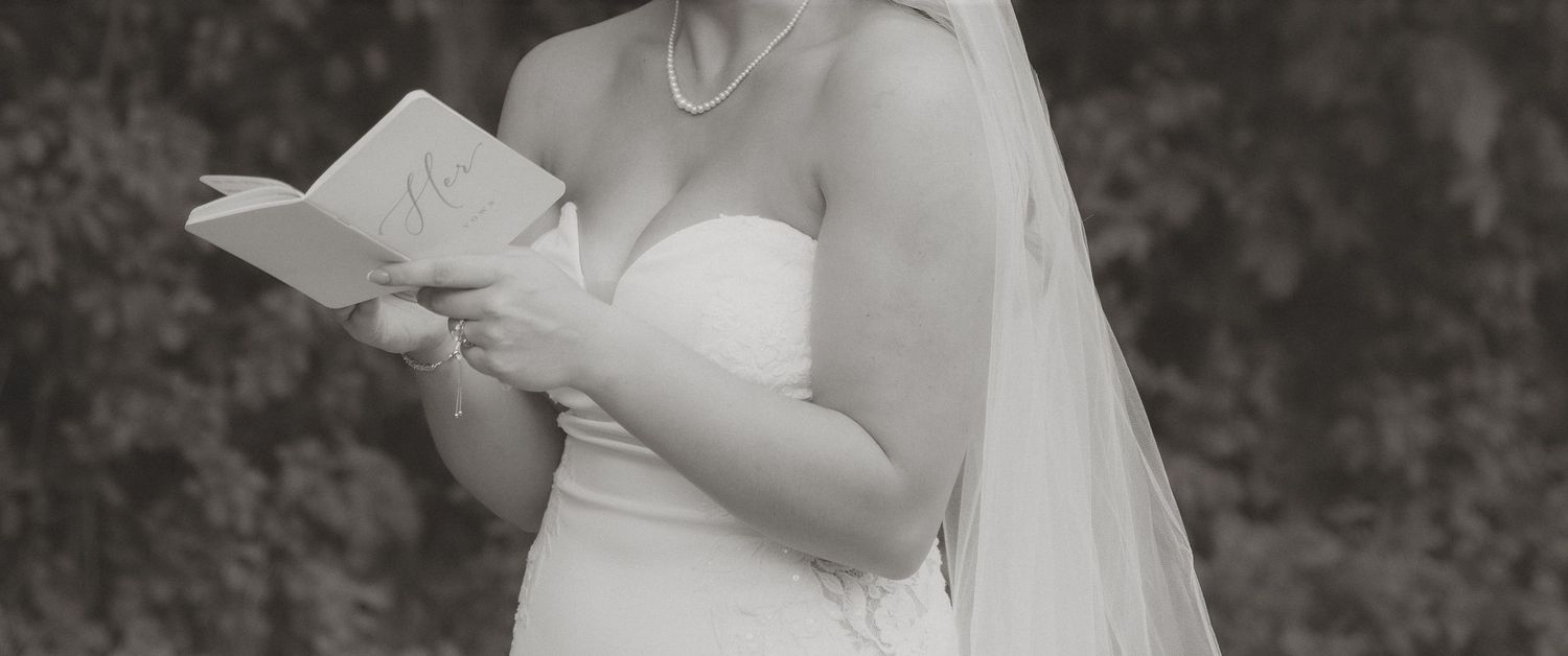 Bride in strapless wedding gown and veil reading vows from a book outdoors in black and white photo.