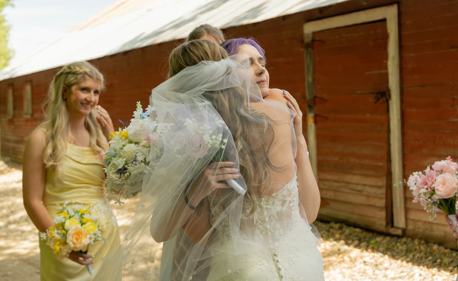 Bride in white gown and veil embracing guest outdoors near red barn with bridesmaid in yellow dress holding bouquet.