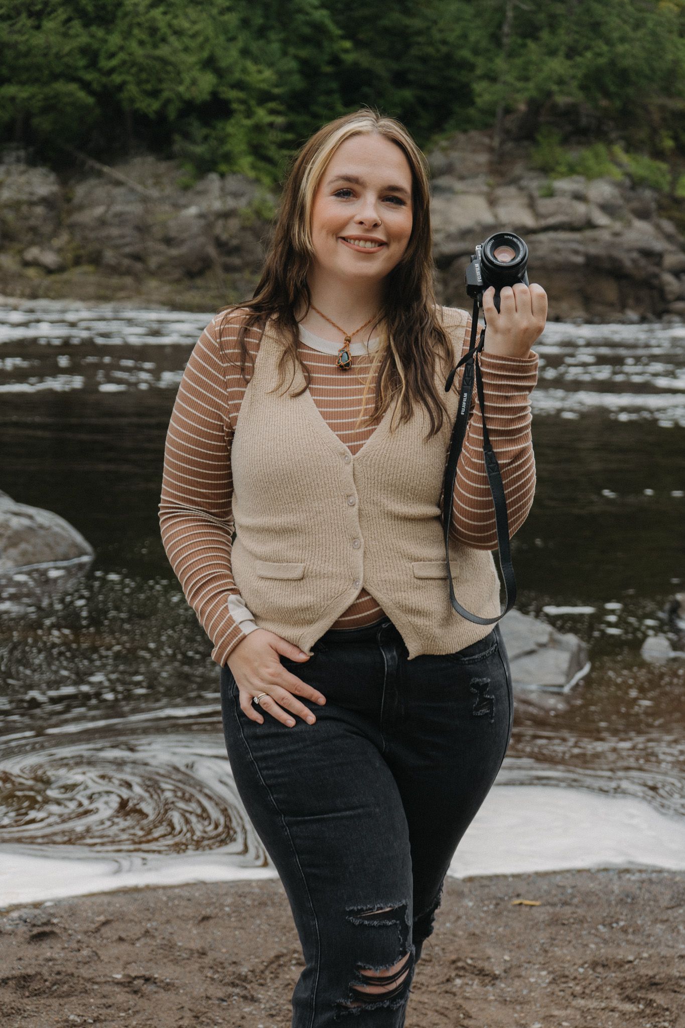Young woman smiling outdoors by a river, holding a camera, wearing a beige vest over a striped shirt and black jeans.