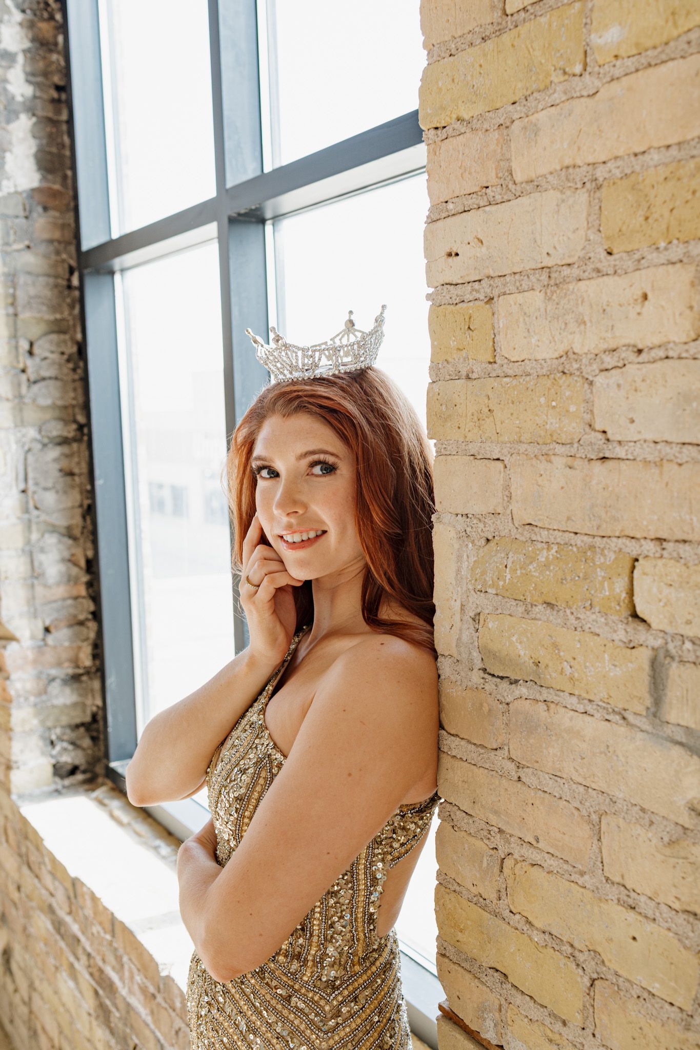 A smiling redhead woman in a gold sequin gown and rhinestone crown poses elegantly by a brick wall near a large window.