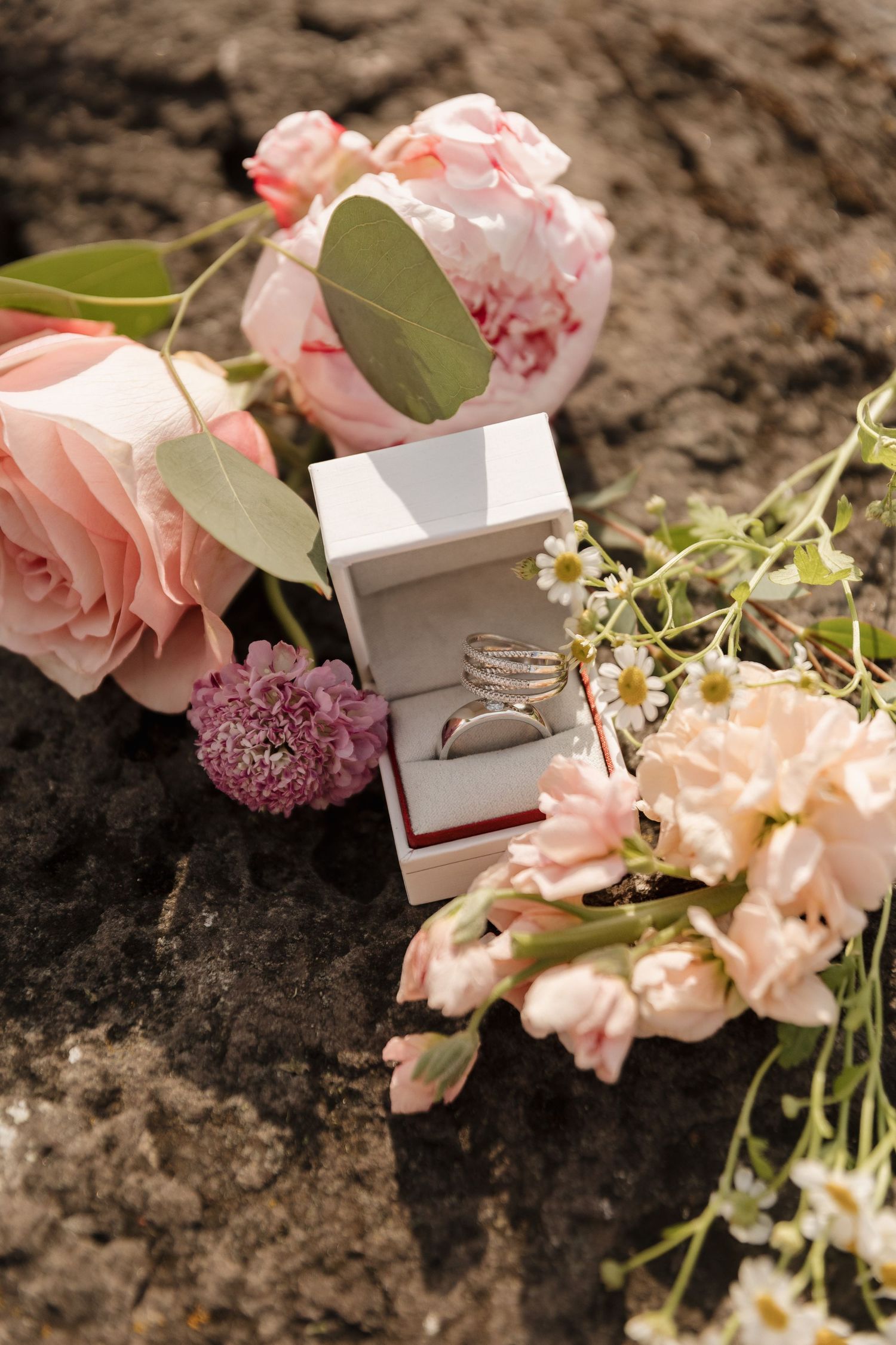 Wedding rings in a white box surrounded by pink roses, peonies, and wildflowers on a rocky surface.