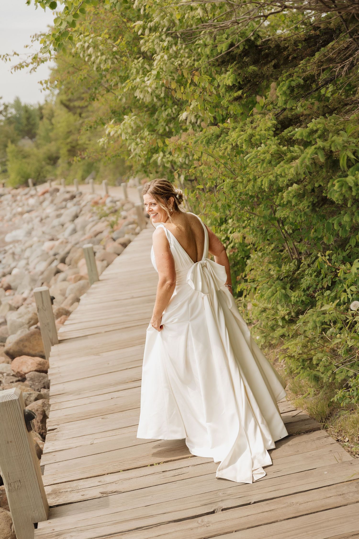Bride in backless white wedding gown with bow walking on wooden boardwalk surrounded by green trees.