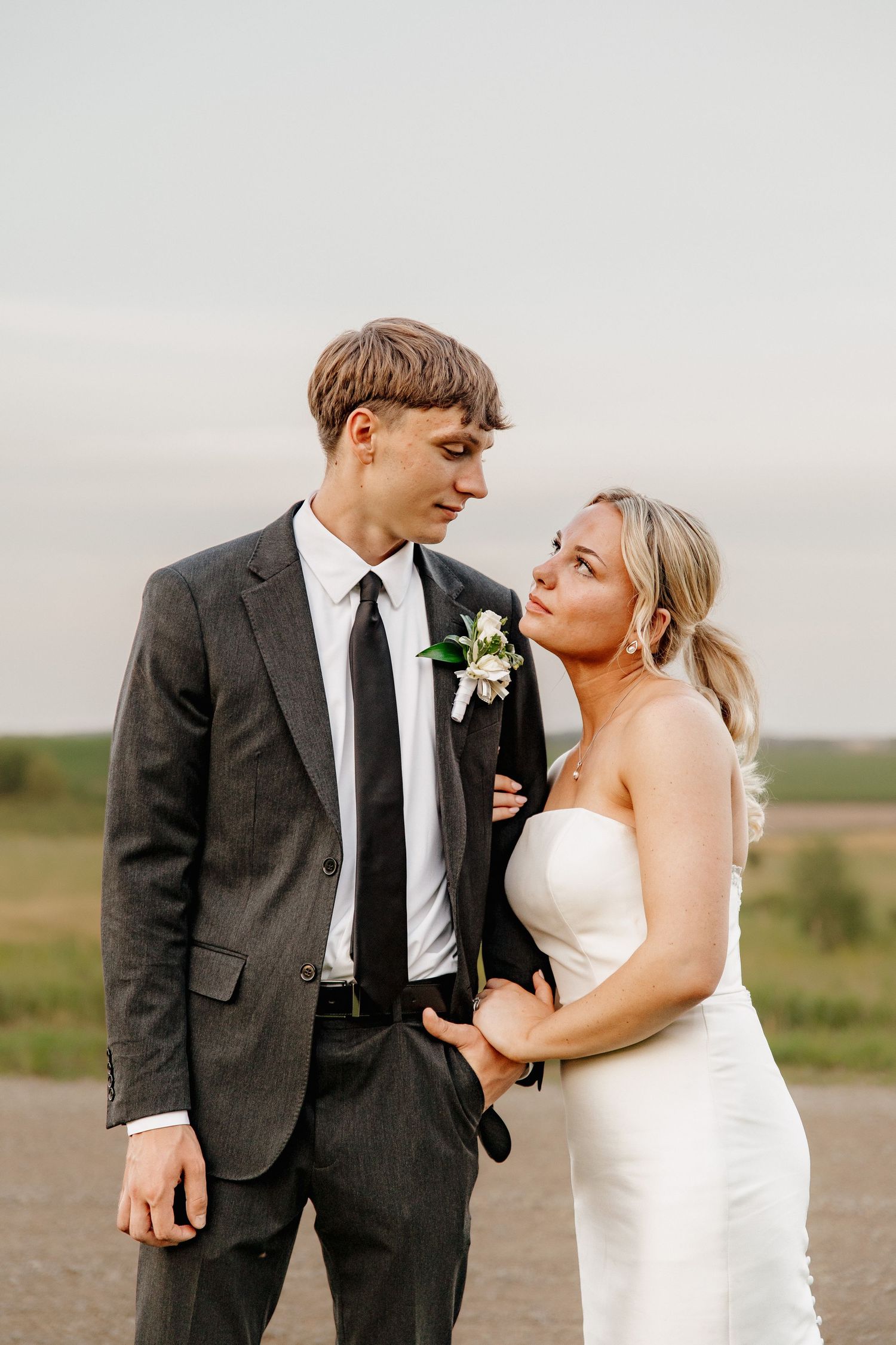 Bride in strapless gown gazing at groom in gray suit with floral boutonniere outdoors.