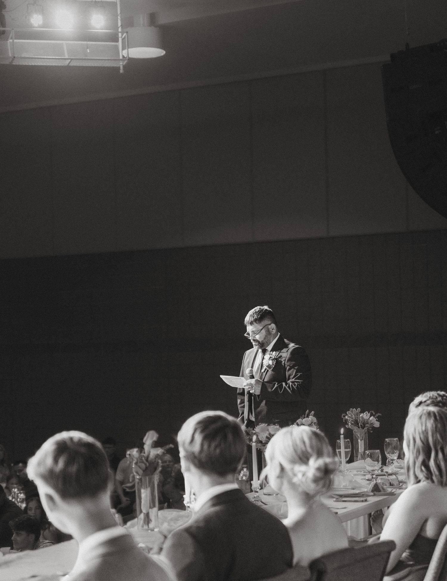 Man delivers heartfelt speech at wedding reception while guests listen at decorated banquet tables.