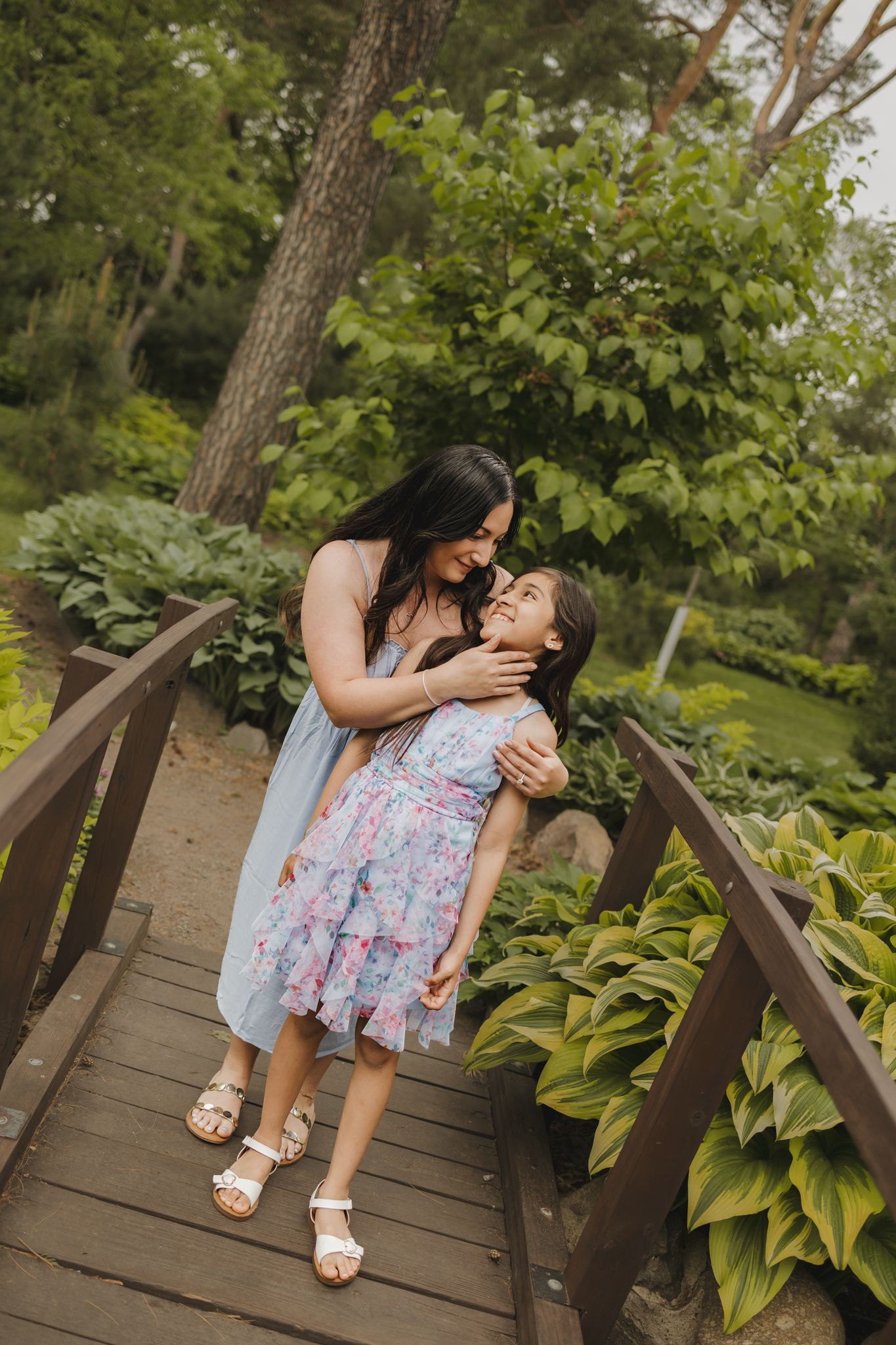 A mother and daughter in floral dresses share a tender embrace on a wooden bridge surrounded by lush green garden foliage.