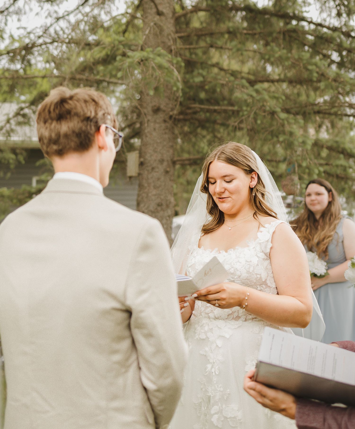 Bride in lace wedding gown reading vows to groom during outdoor wedding ceremony near pine tree.
