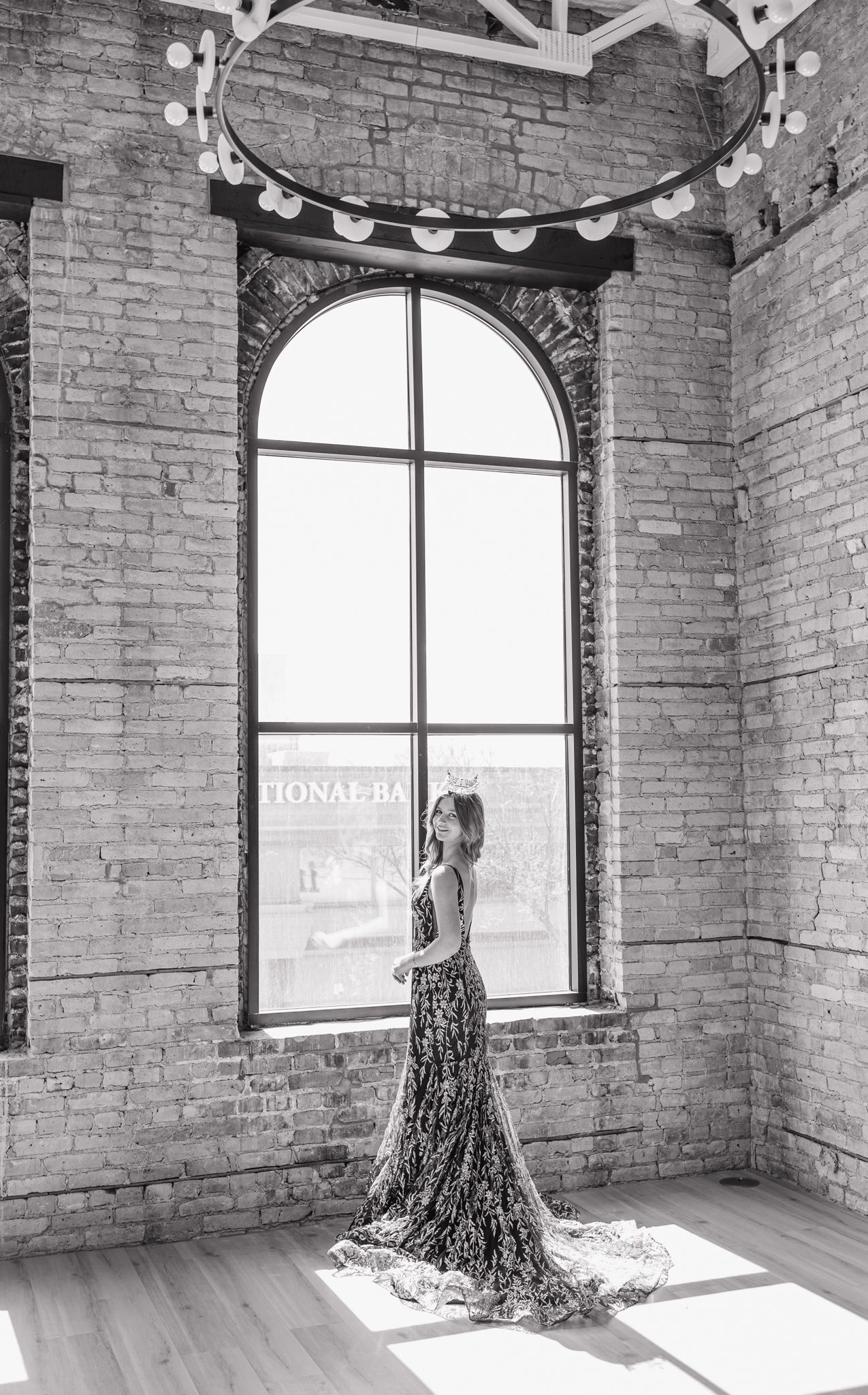 Woman in floral gown posing by tall arched window in black and white loft photo.