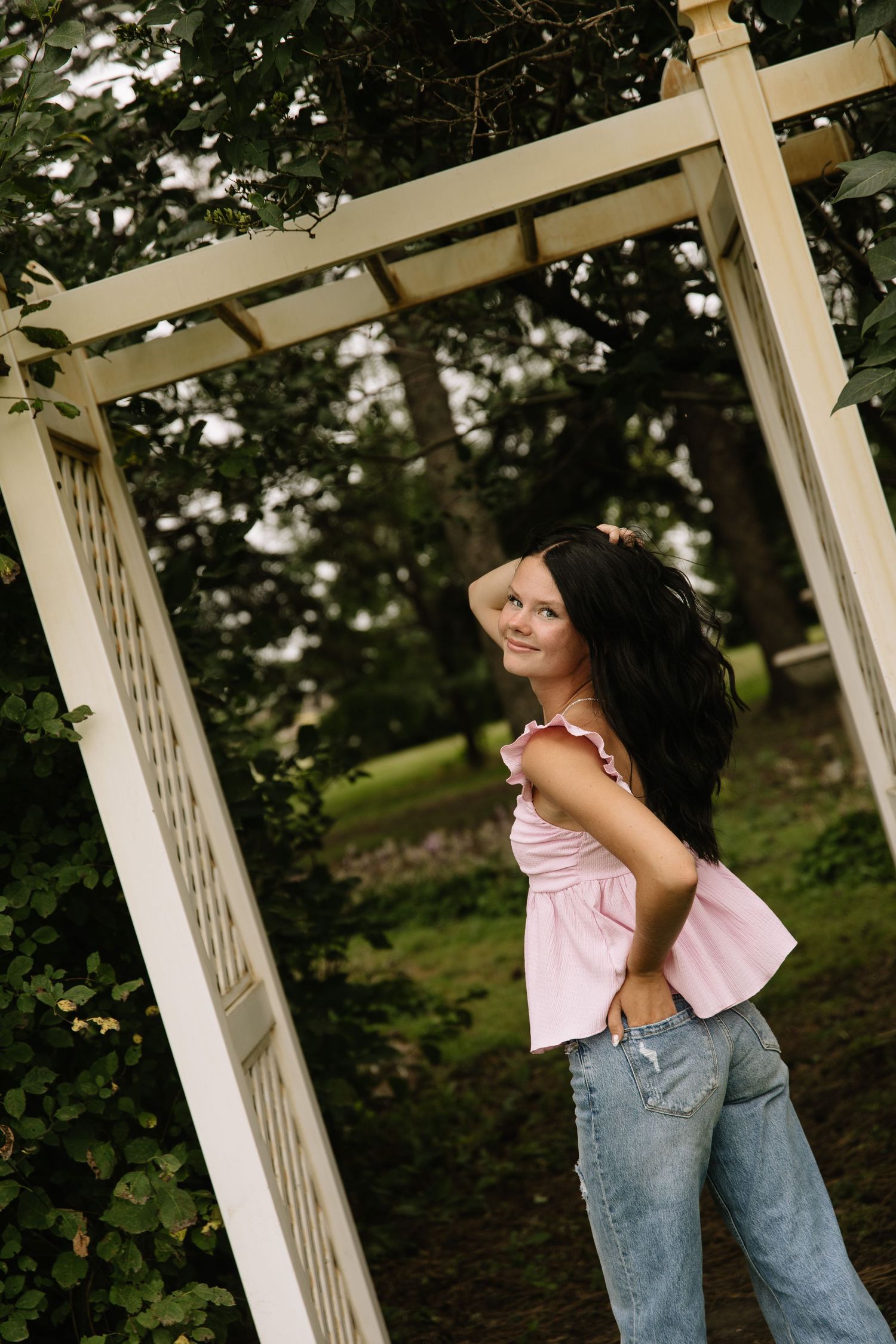 Young woman in pink top and jeans posing under a white garden arbor surrounded by lush green trees.