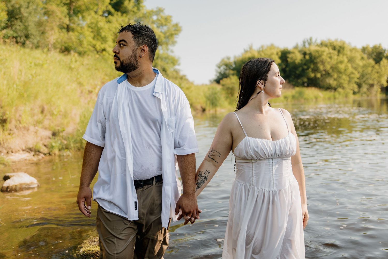 Couple holding hands standing in a shallow river, wearing white outfits, surrounded by lush green trees.