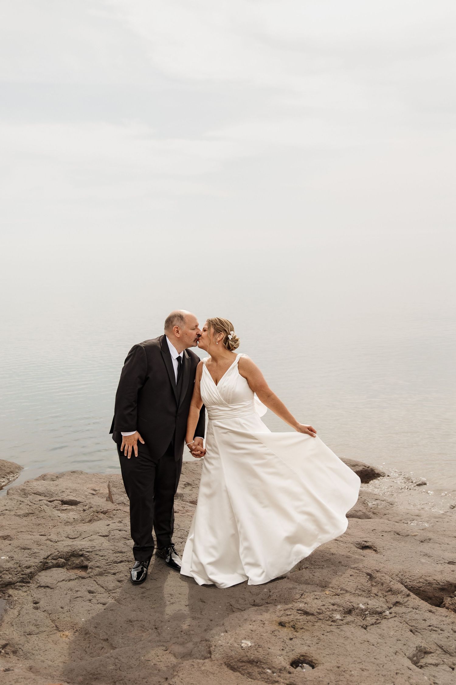 Bride and groom kissing on rocky shoreline, white gown flowing in breeze, overcast sky and calm water backdrop.
