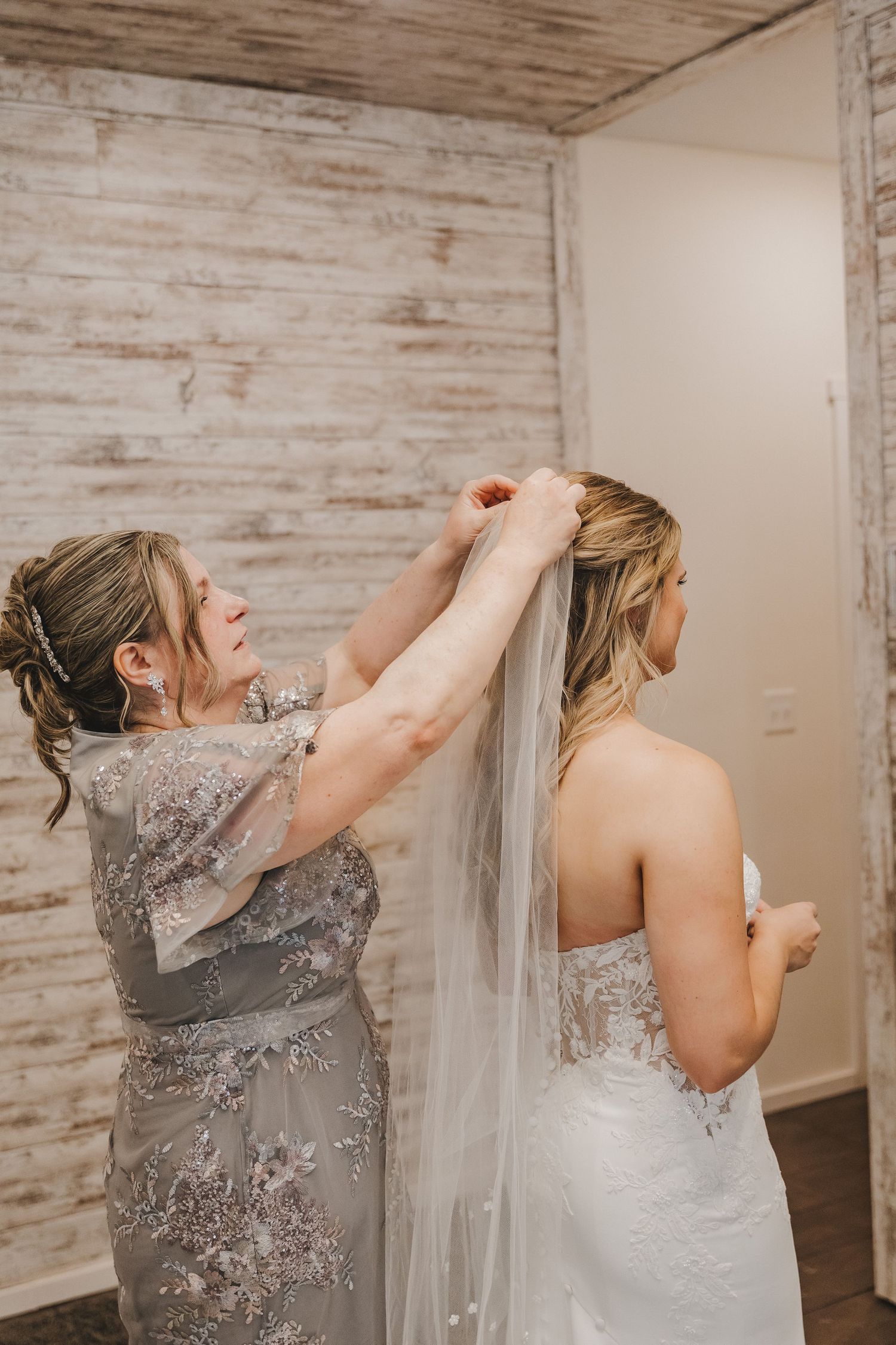 A woman in a gray floral gown helps a bride attach her veil before the wedding ceremony.
