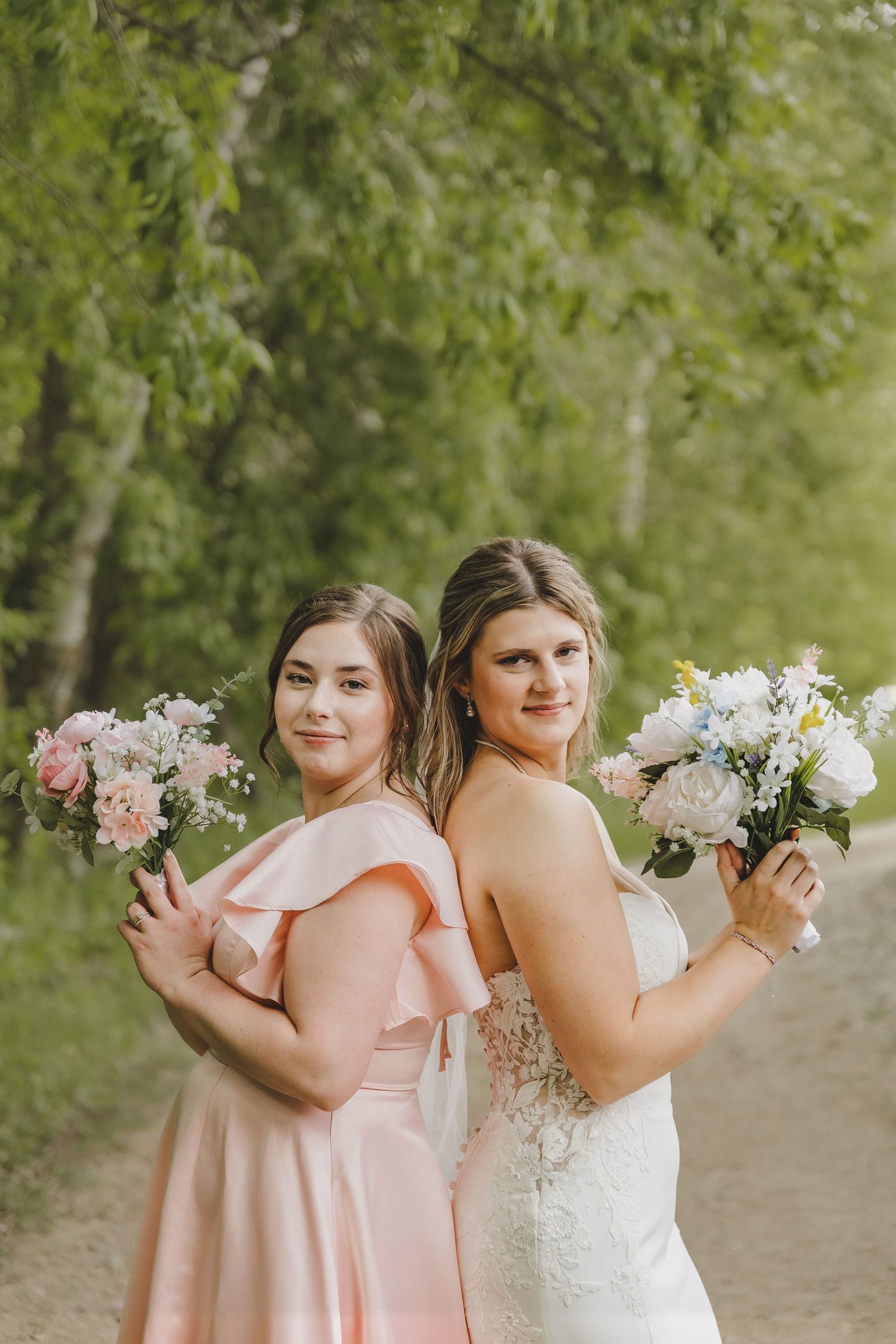 A bride in a lace gown and bridesmaid in a pink dress pose back-to-back holding floral bouquets outdoors.
