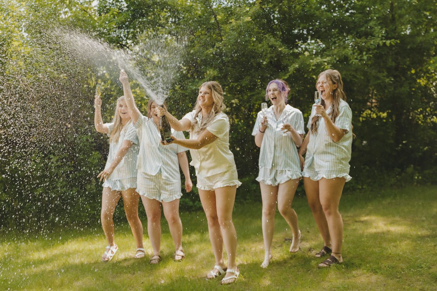 Five women in matching white outfits celebrate outdoors, laughing as champagne sprays in a sunny green backyard.