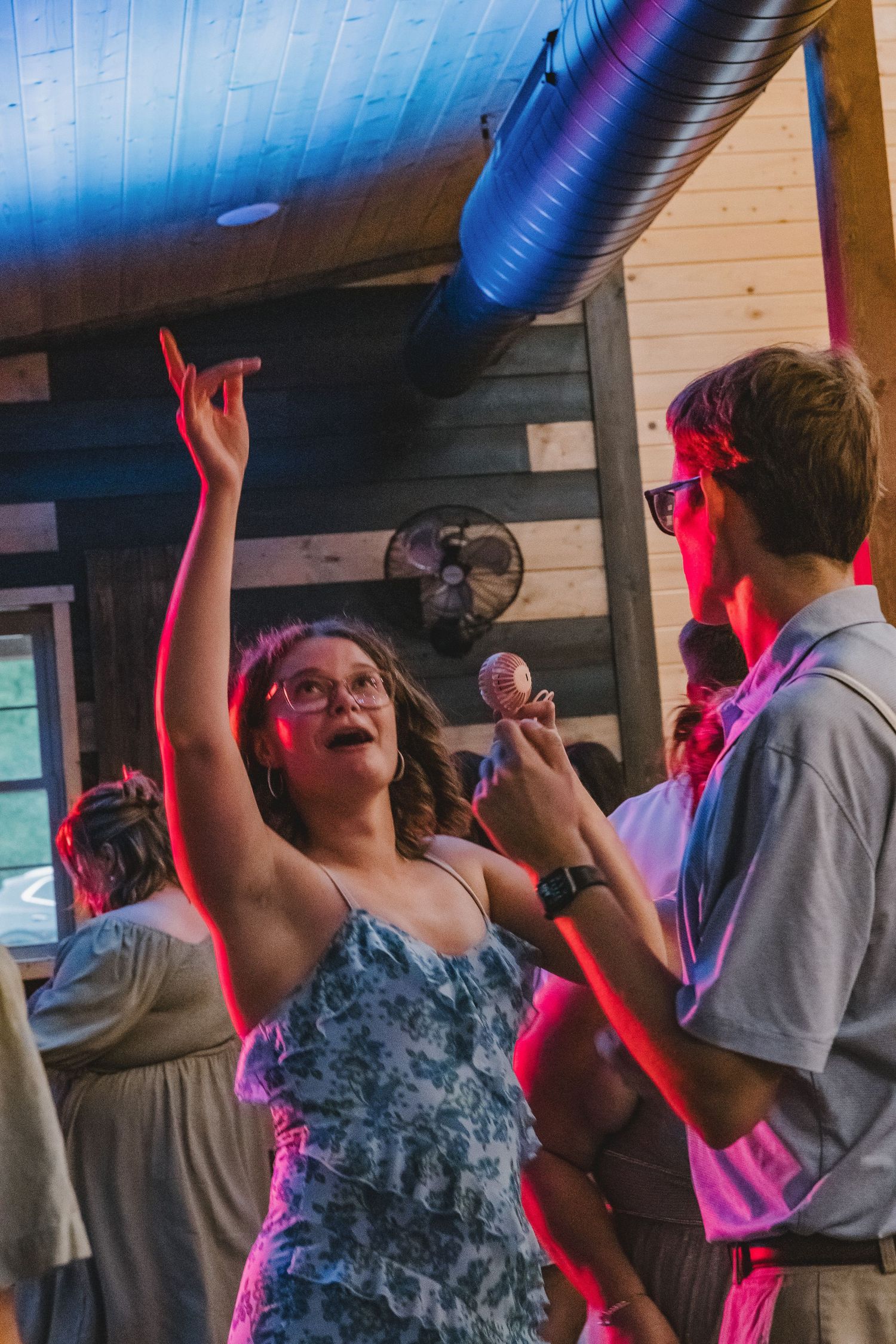 People dancing at a party in a rustic log cabin venue with colorful pink and red lighting.
