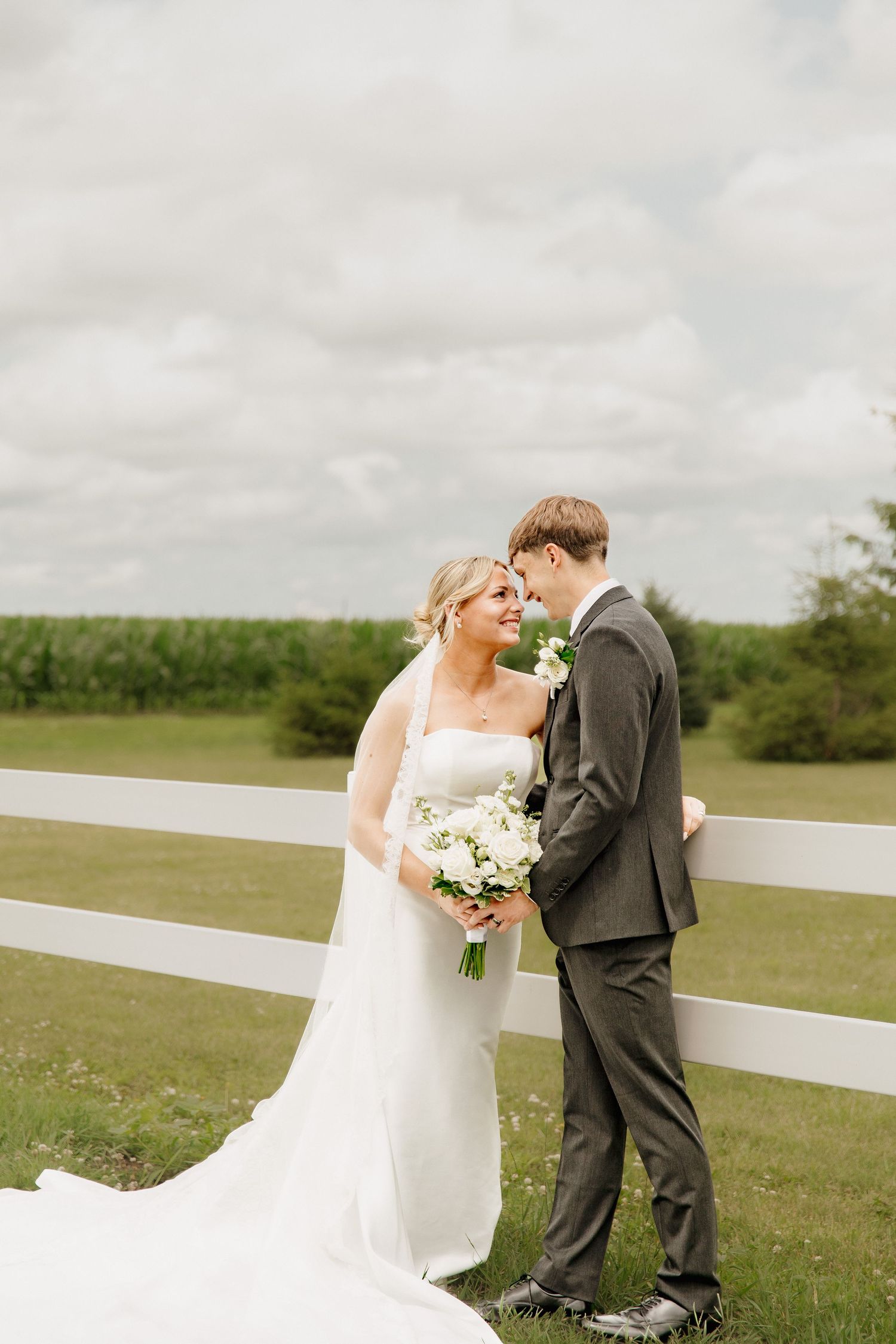 Bride and groom share a romantic moment by a white fence in a green countryside setting.