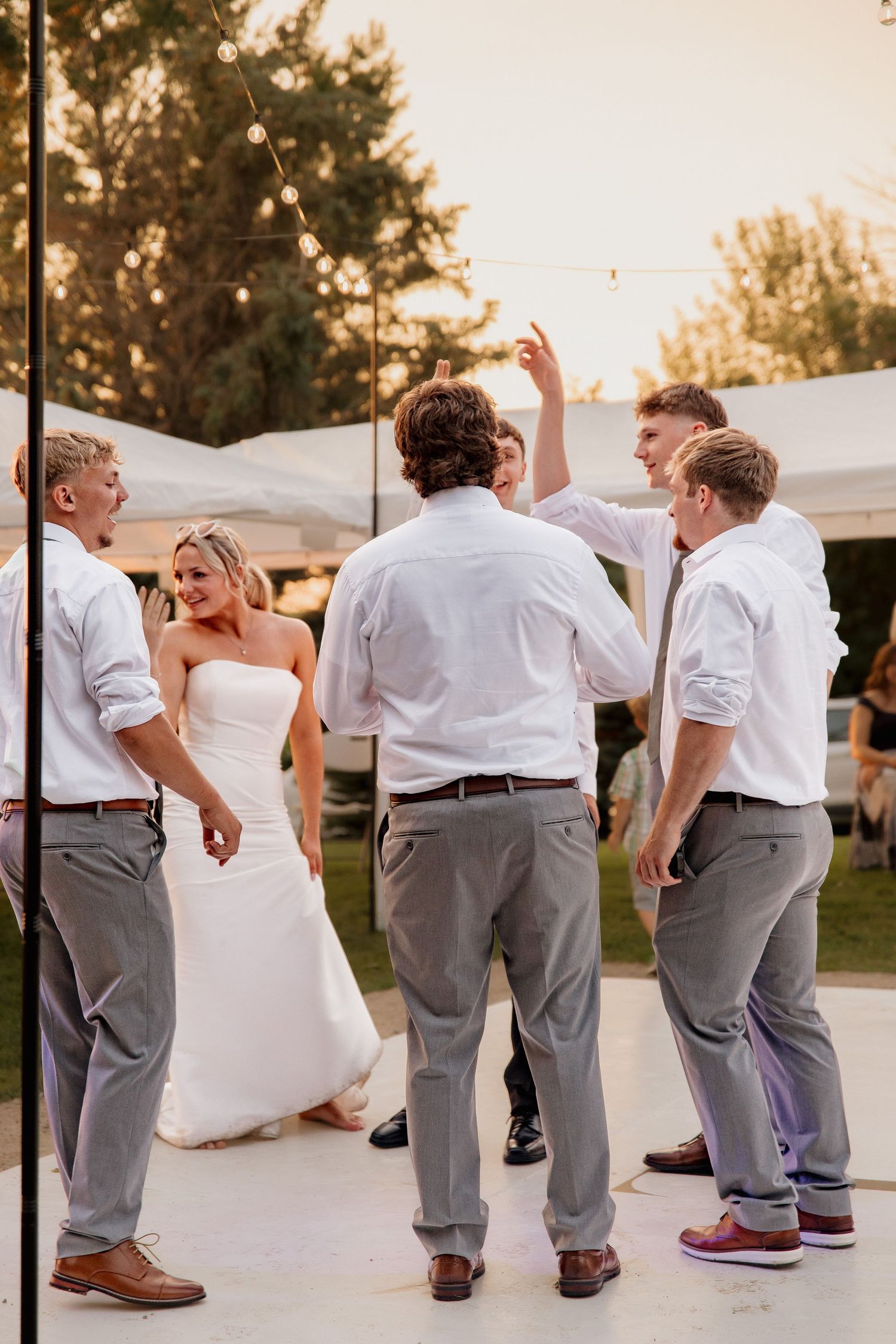 Bride in white strapless gown dances joyfully with groomsmen on outdoor dance floor at golden hour reception.