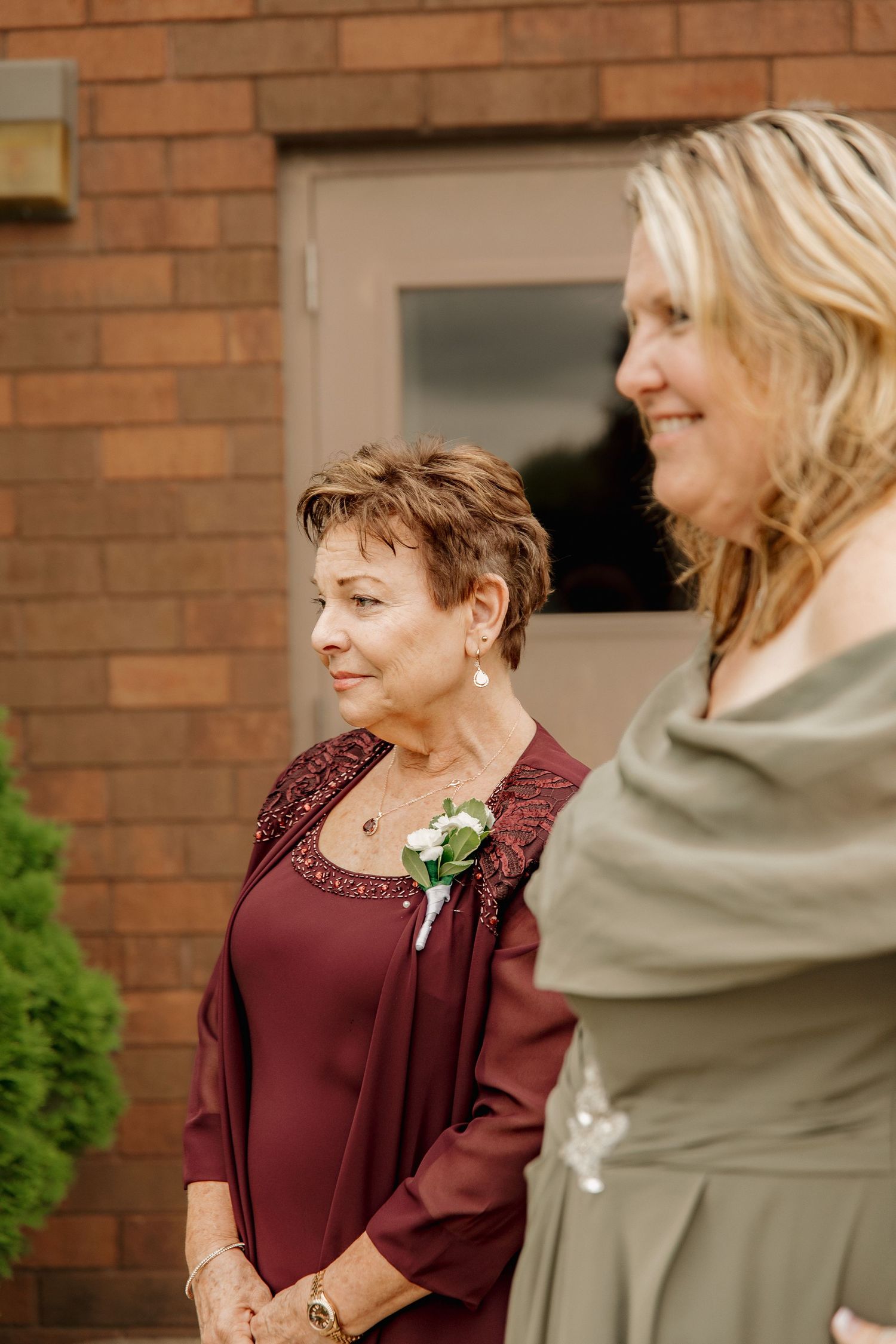 Two elegantly dressed women at a wedding, one in burgundy with a boutonniere, stand together outside a brick building.