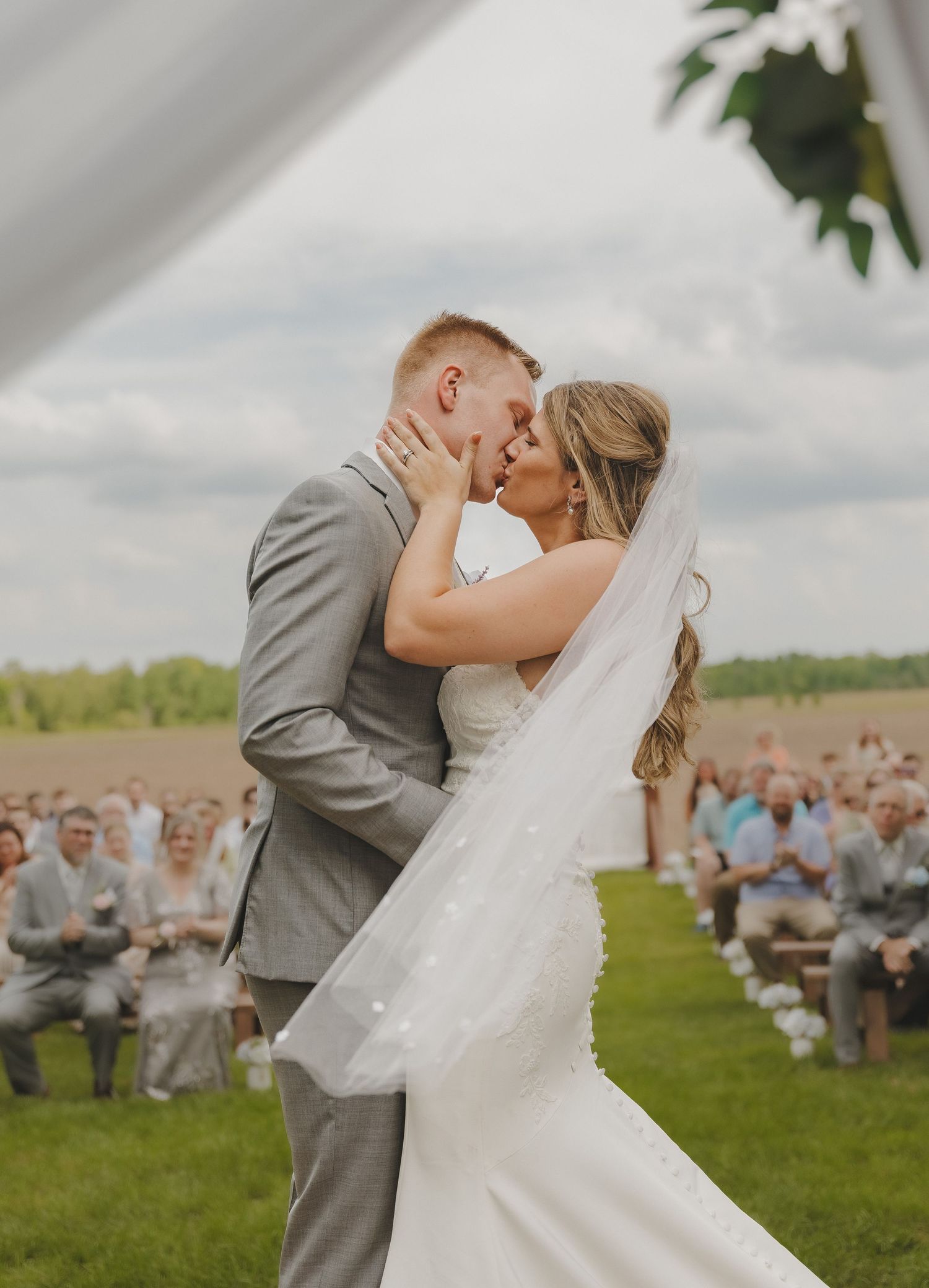 A bride and groom share their first kiss at an outdoor wedding ceremony with guests seated in the background.