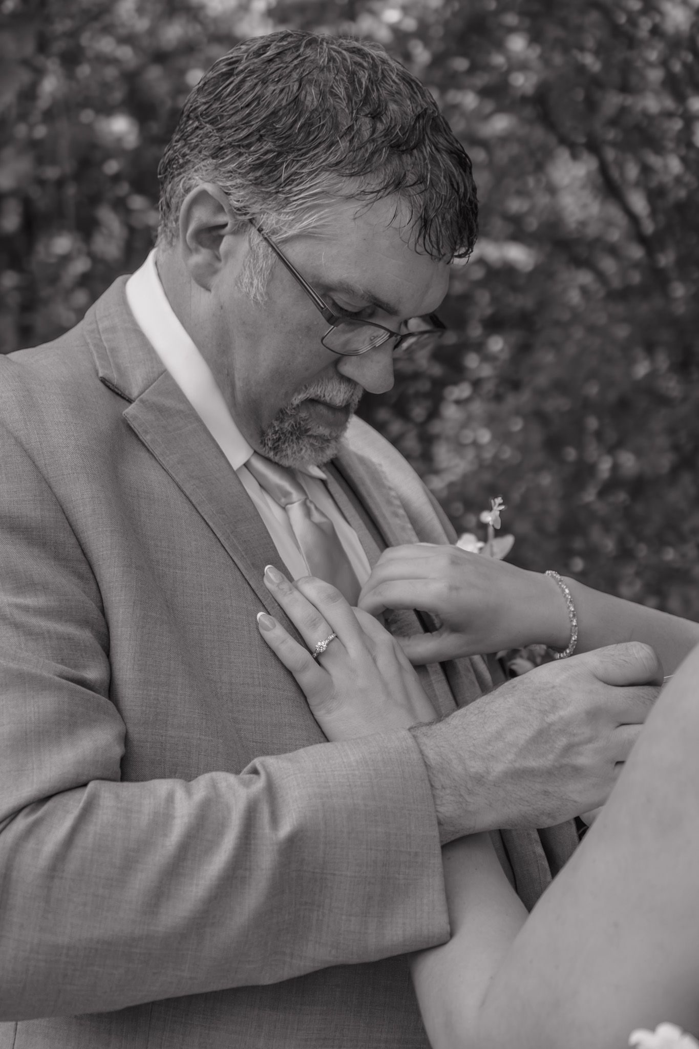Black and white photo of groom in gray suit adjusting boutonniere with help from bride at outdoor wedding ceremony.