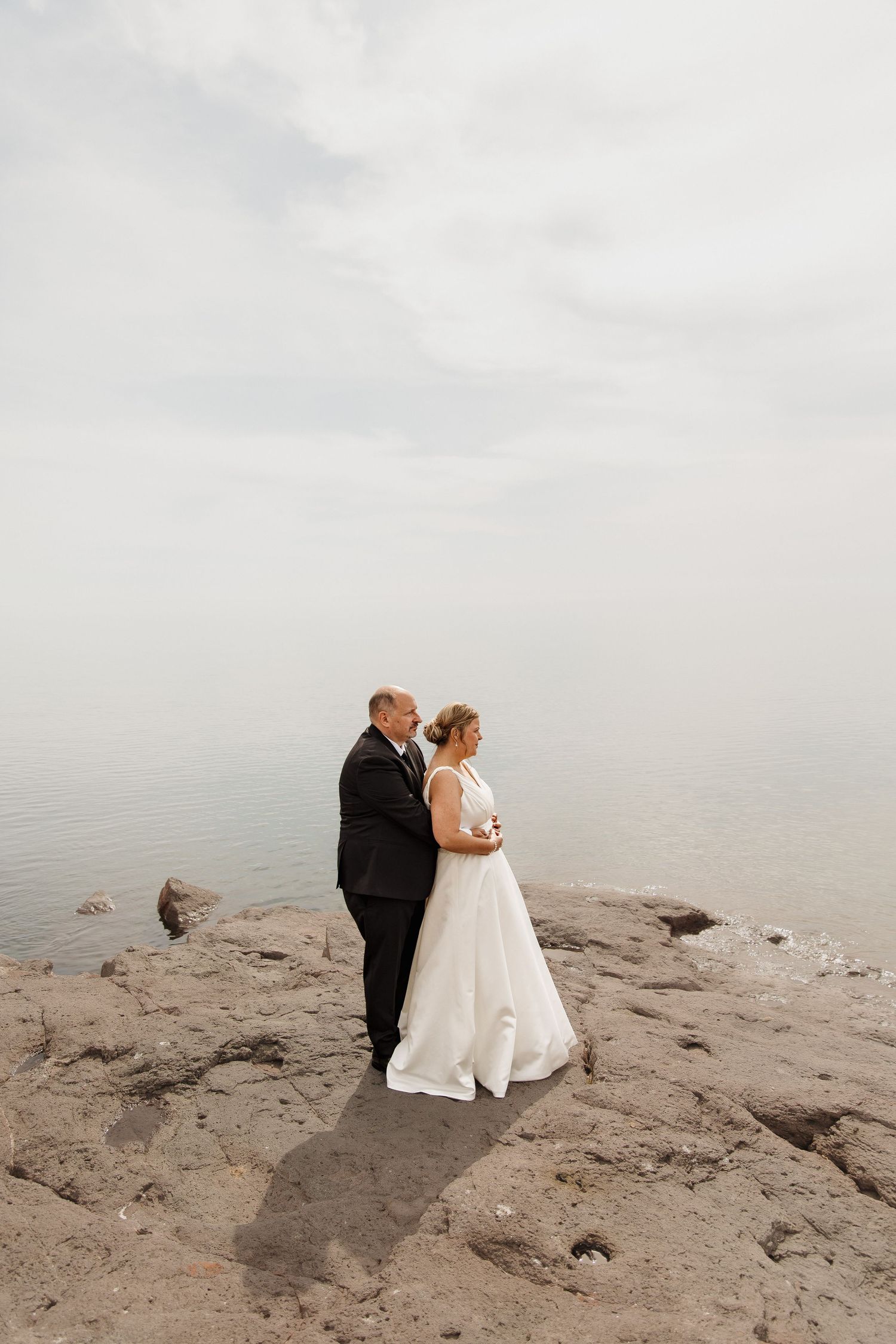 A bride and groom stand on rocky shoreline in wedding attire, with calm misty water and overcast sky behind them.