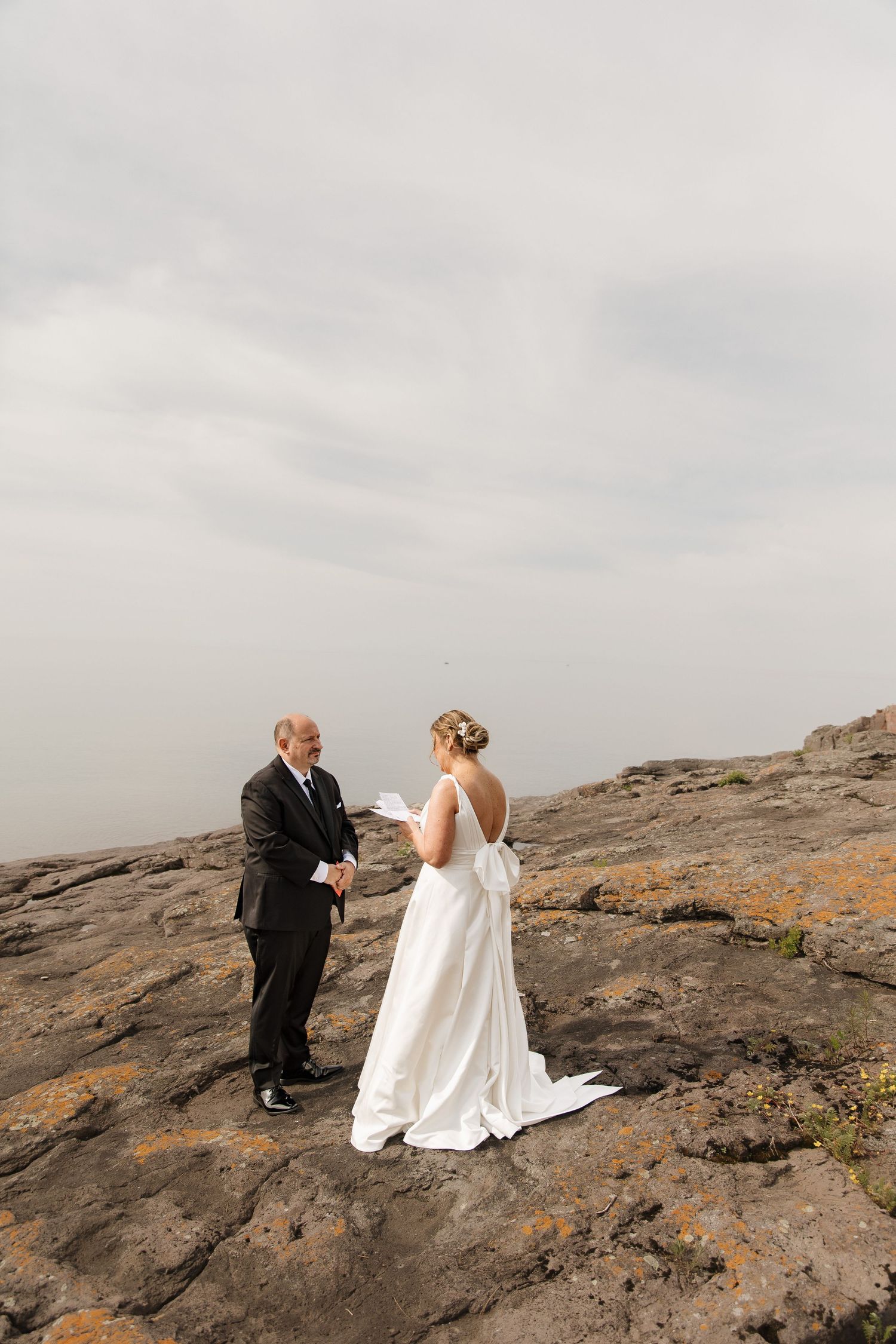 Bride and groom exchange vows on rocky mountaintop, bride in white gown holding paper, groom in black suit, overcast sky.