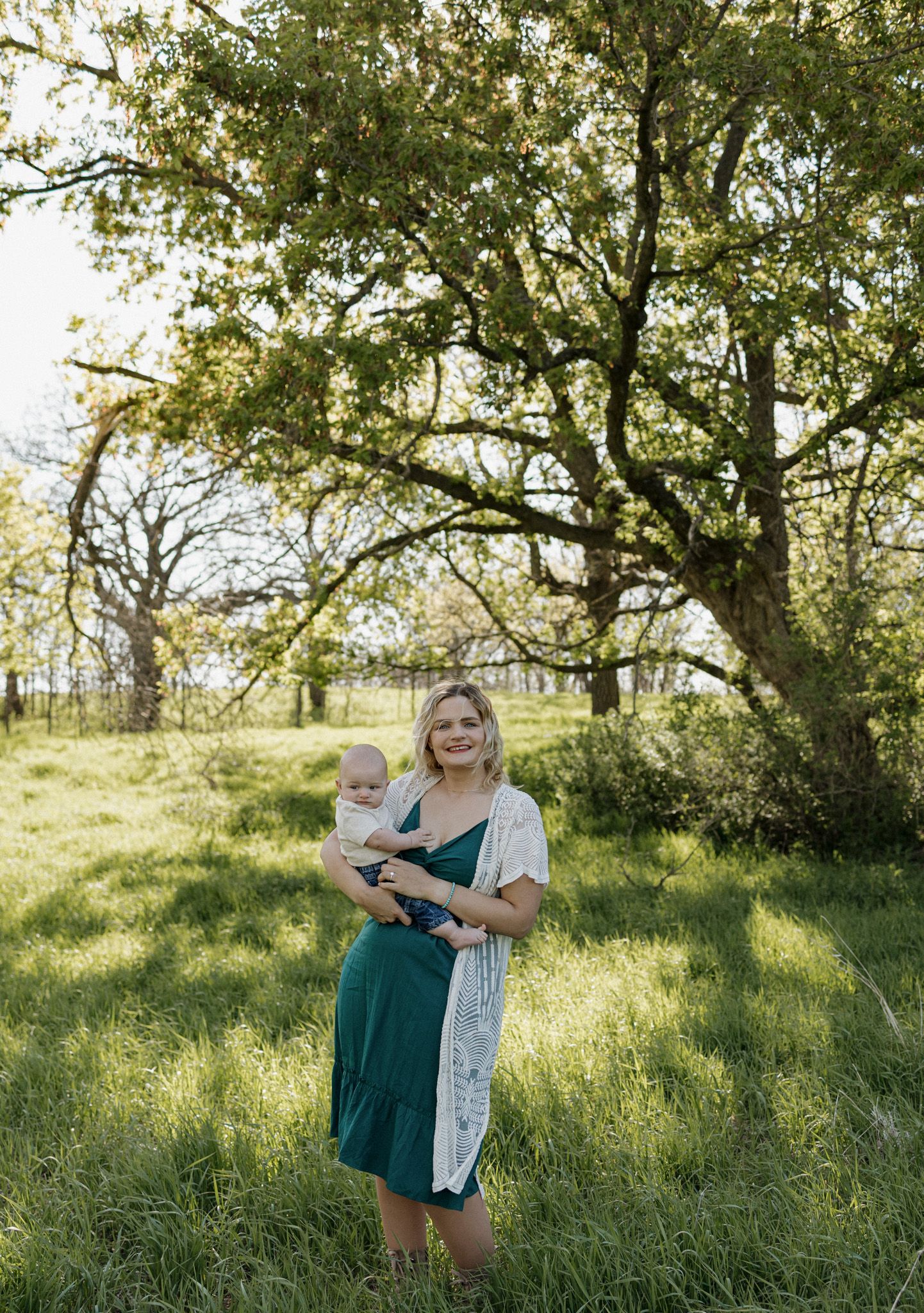 Smiling woman in green dress holding baby outdoors in lush green field with large oak tree background.