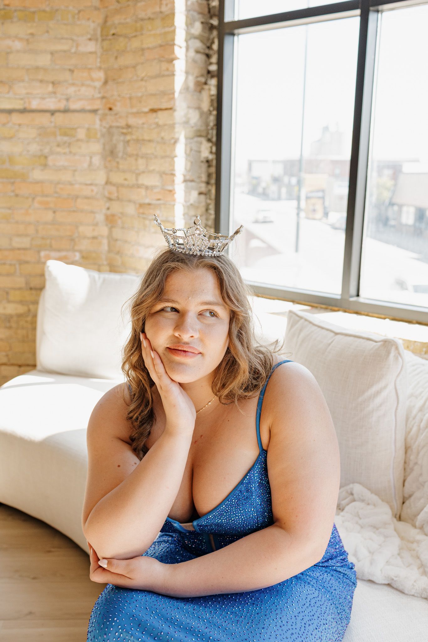 Woman wearing a blue sequin dress and tiara sits thoughtfully on a white couch near a sunny window.