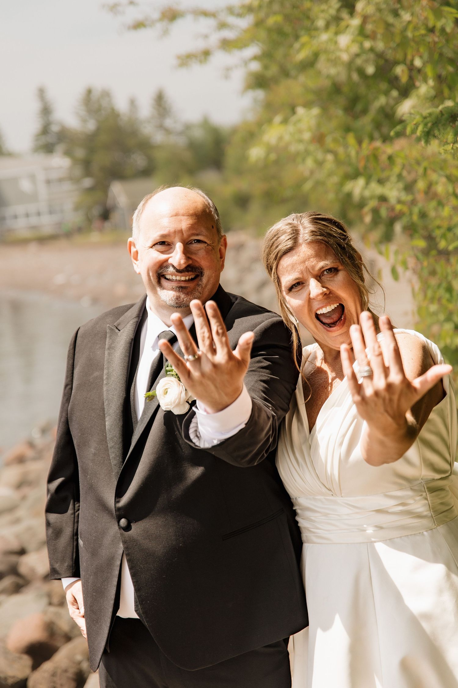 Joyful newlyweds showing off wedding rings to camera near a scenic lakeside setting.