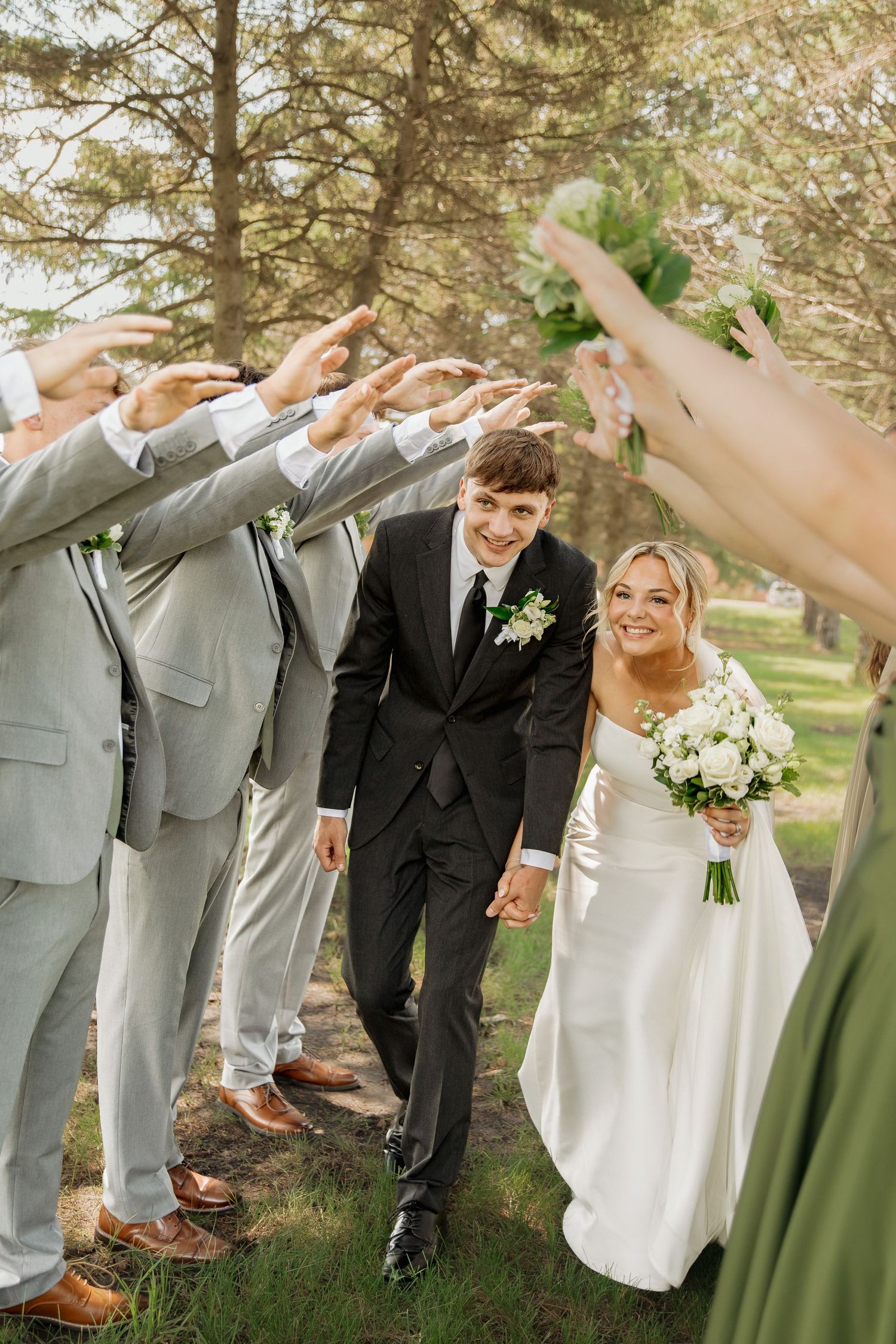 Bride and groom walk hand-in-hand through an arch of hands formed by groomsmen in gray suits outdoors.