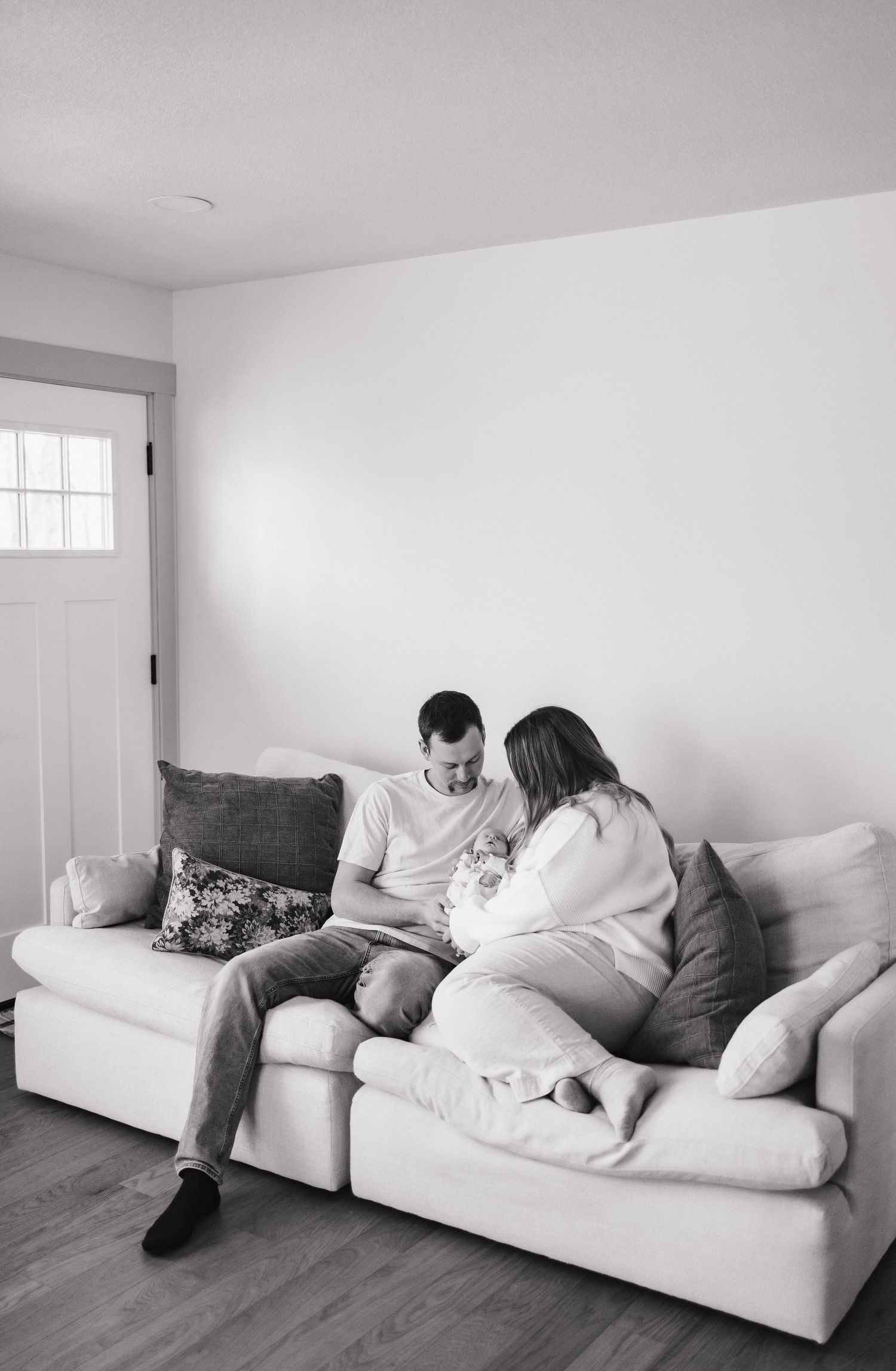 Black and white photo of a couple sitting on a cozy sofa, looking down at their newborn baby together.