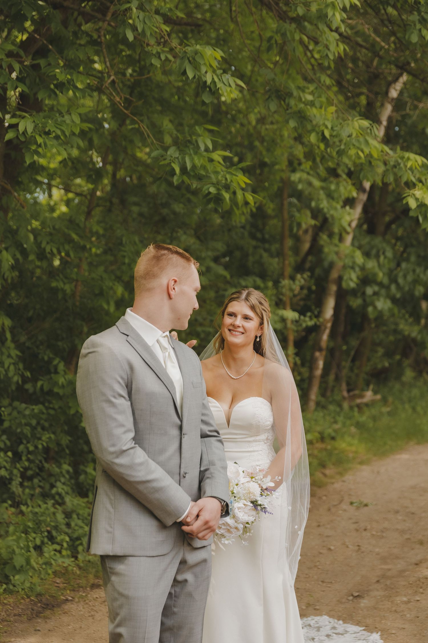 Bride in a strapless white gown with veil smiles at groom in gray suit on a tree-lined path.