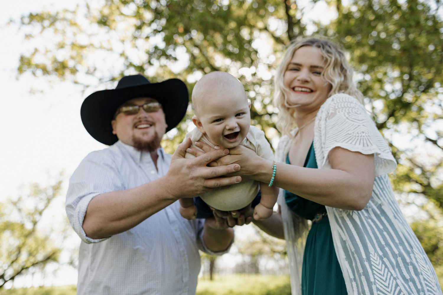 Happy family outdoors holding laughing baby, man in cowboy hat and woman in green dress, trees in background.