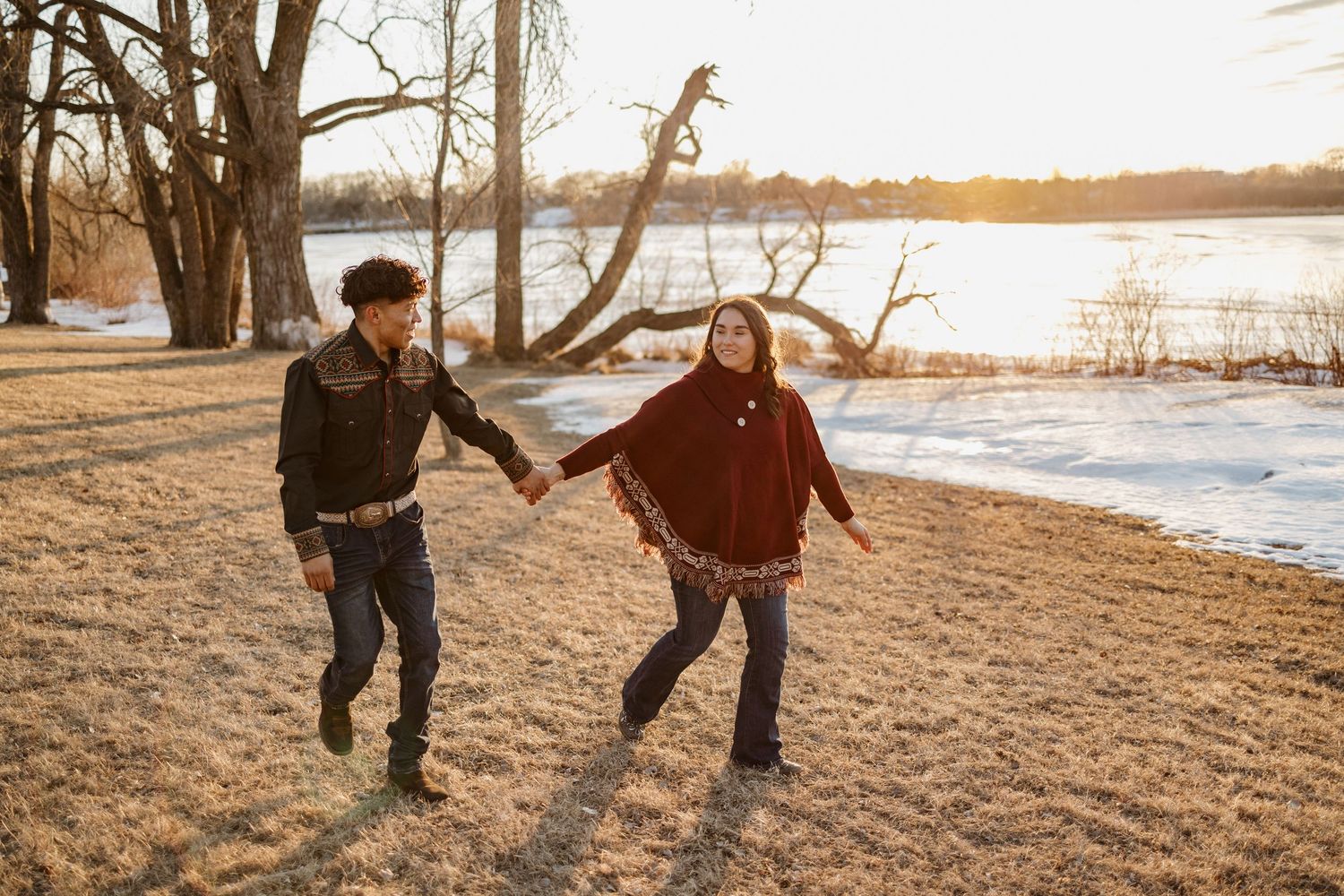 Two people playfully run together along a snowy lakeshore at sunset with bare trees in the background.