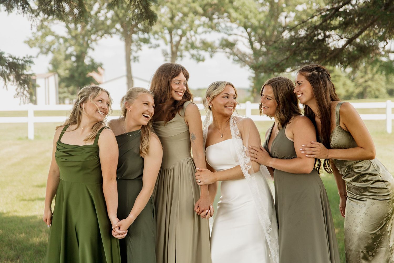 Bride in white gown laughing with five bridesmaids in sage green dresses outdoors at a wedding.