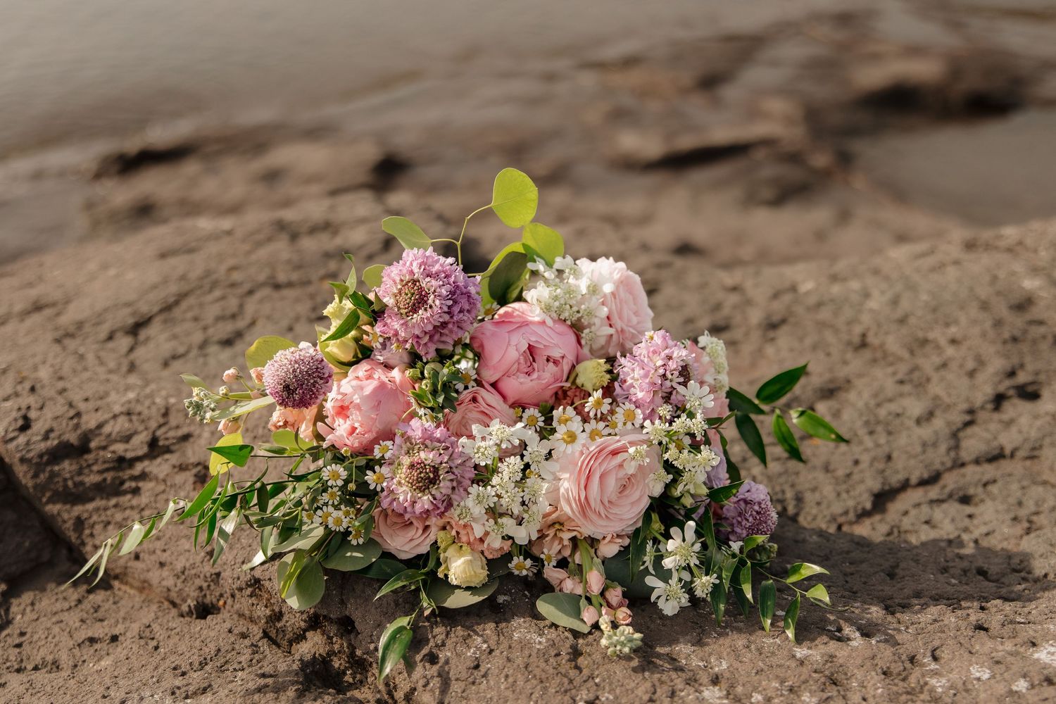Pink and purple floral bouquet with roses and wildflowers resting on sandy rocks near water.