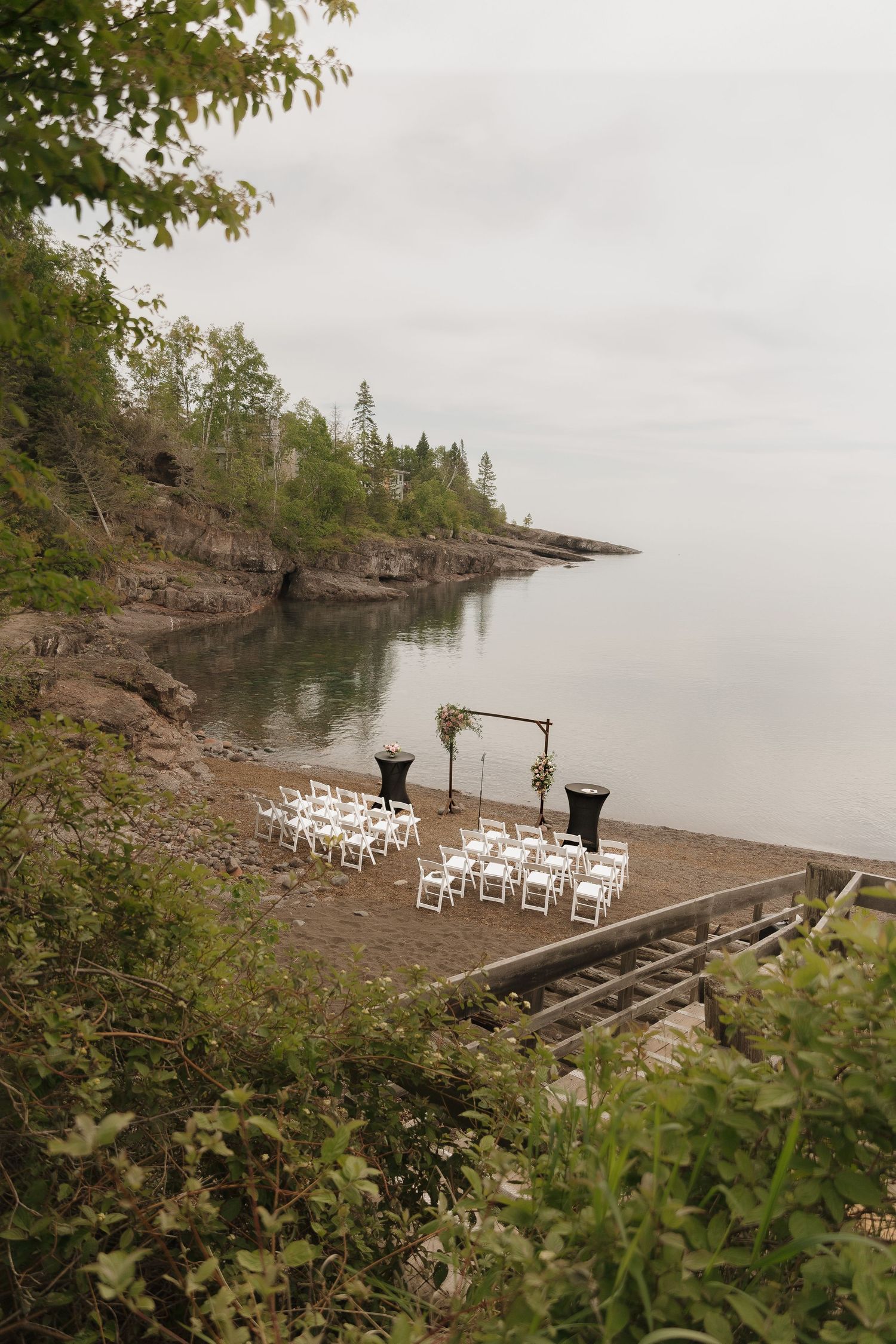 An intimate outdoor wedding ceremony setup on a rocky lakeside beach surrounded by lush green trees.