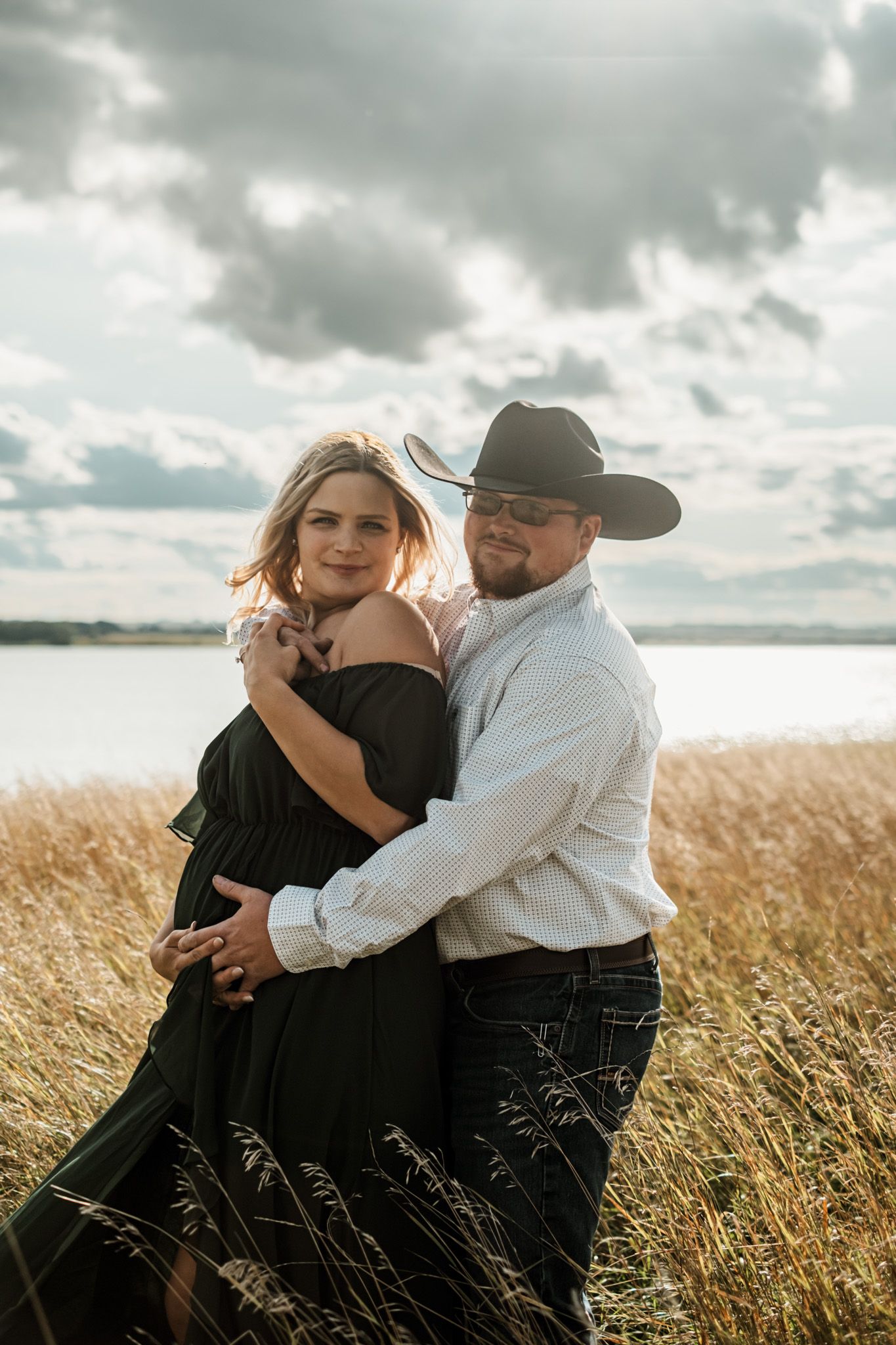 Intimate couple's portrait in black dress and western wear among wheat field at sunset.