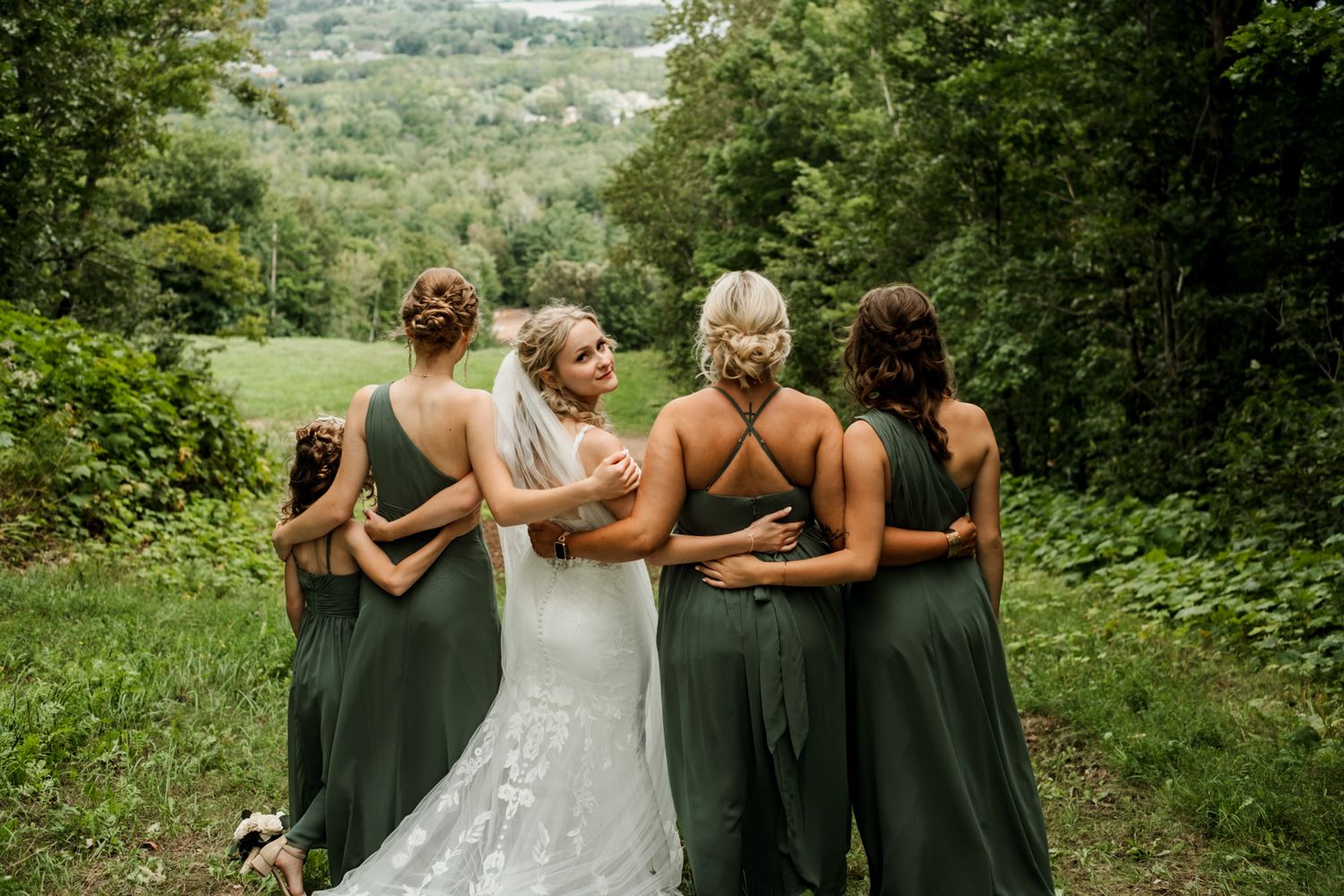 Bridesmaids in dark green dresses pose with bride for group photo outdoors.