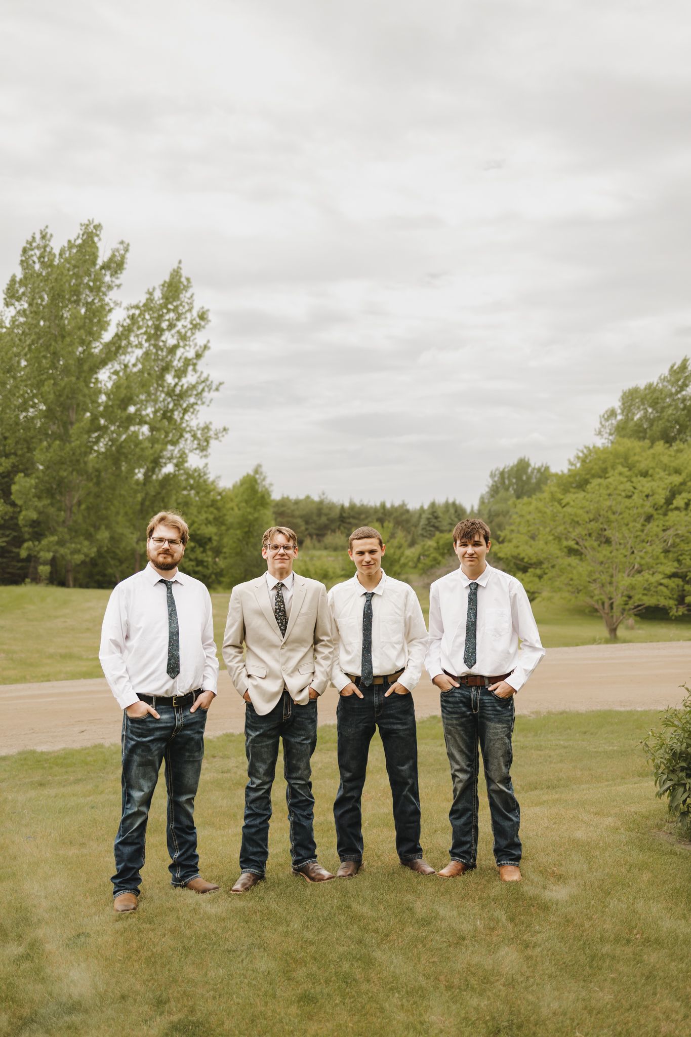 Four groomsmen in white shirts, ties, and jeans pose outdoors on a green lawn with trees in the background.