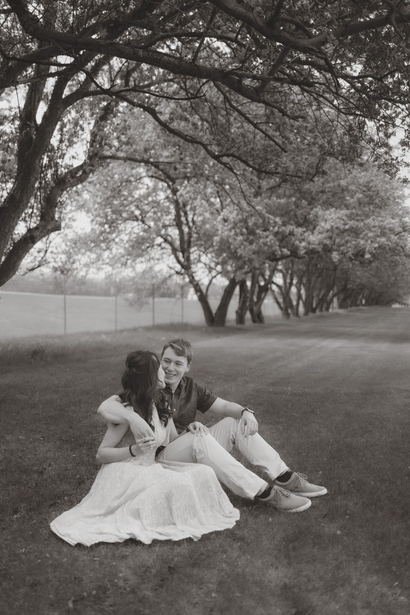 Black and white photo of a couple sitting together on grass under trees in a park, sharing a tender moment.